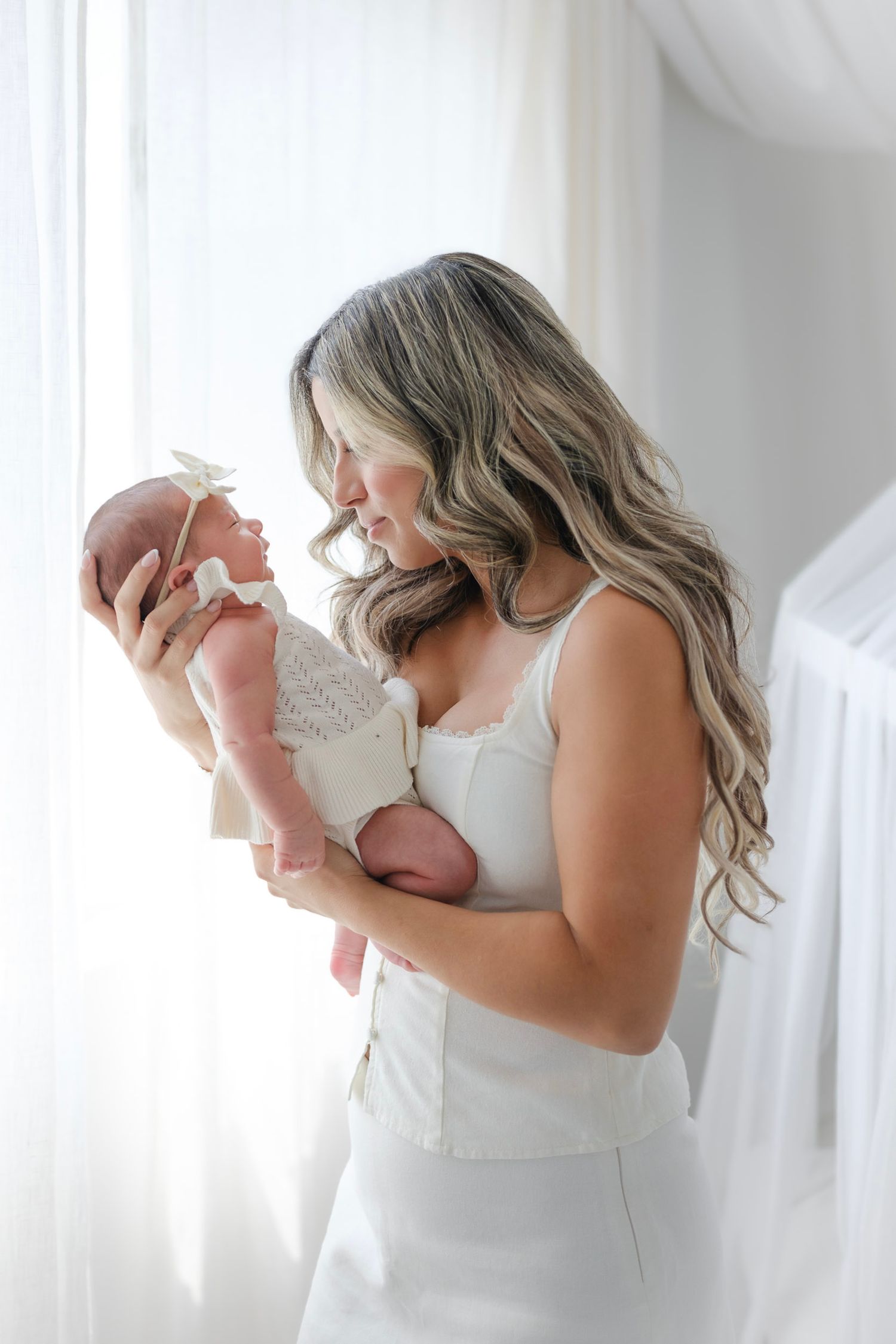 New mother in white dress holding her newborn baby near a bright window, gazing lovingly at her child.