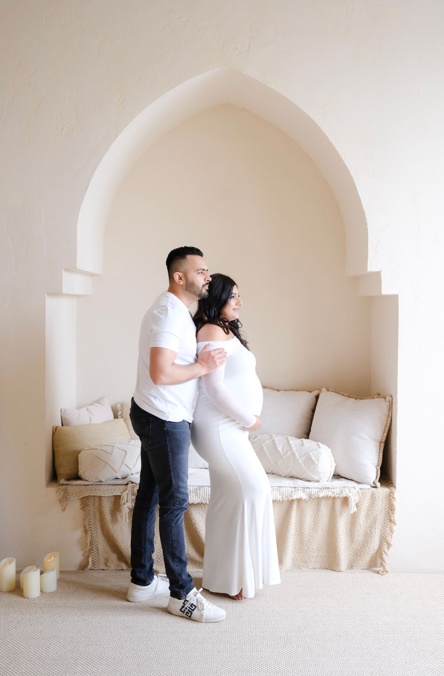A couple embraces in an elegant white arched alcove during a maternity photoshoot with candles and cushions.