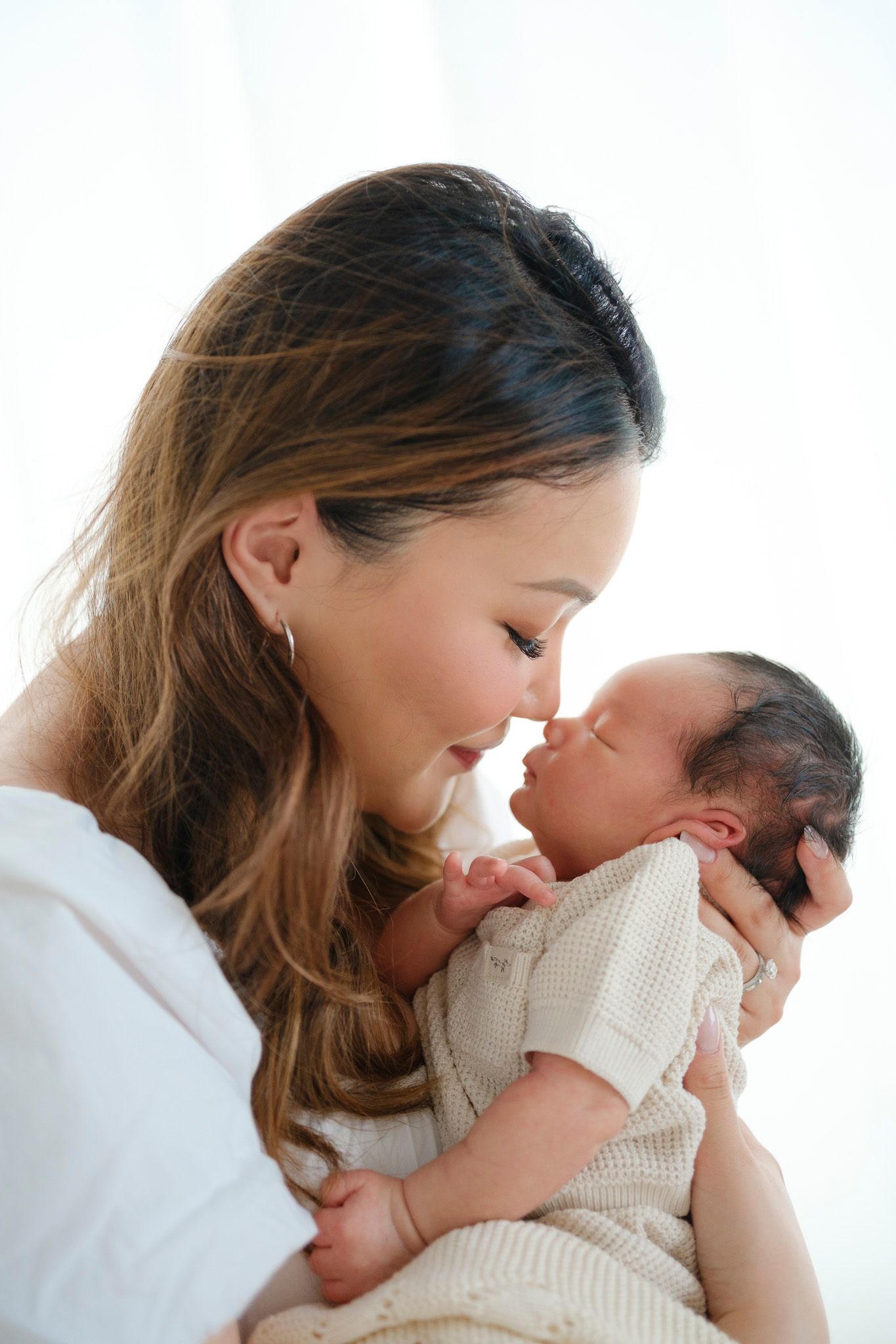 A mother sharing a tender nose-to-nose moment with her newborn baby wearing a cream knit outfit.