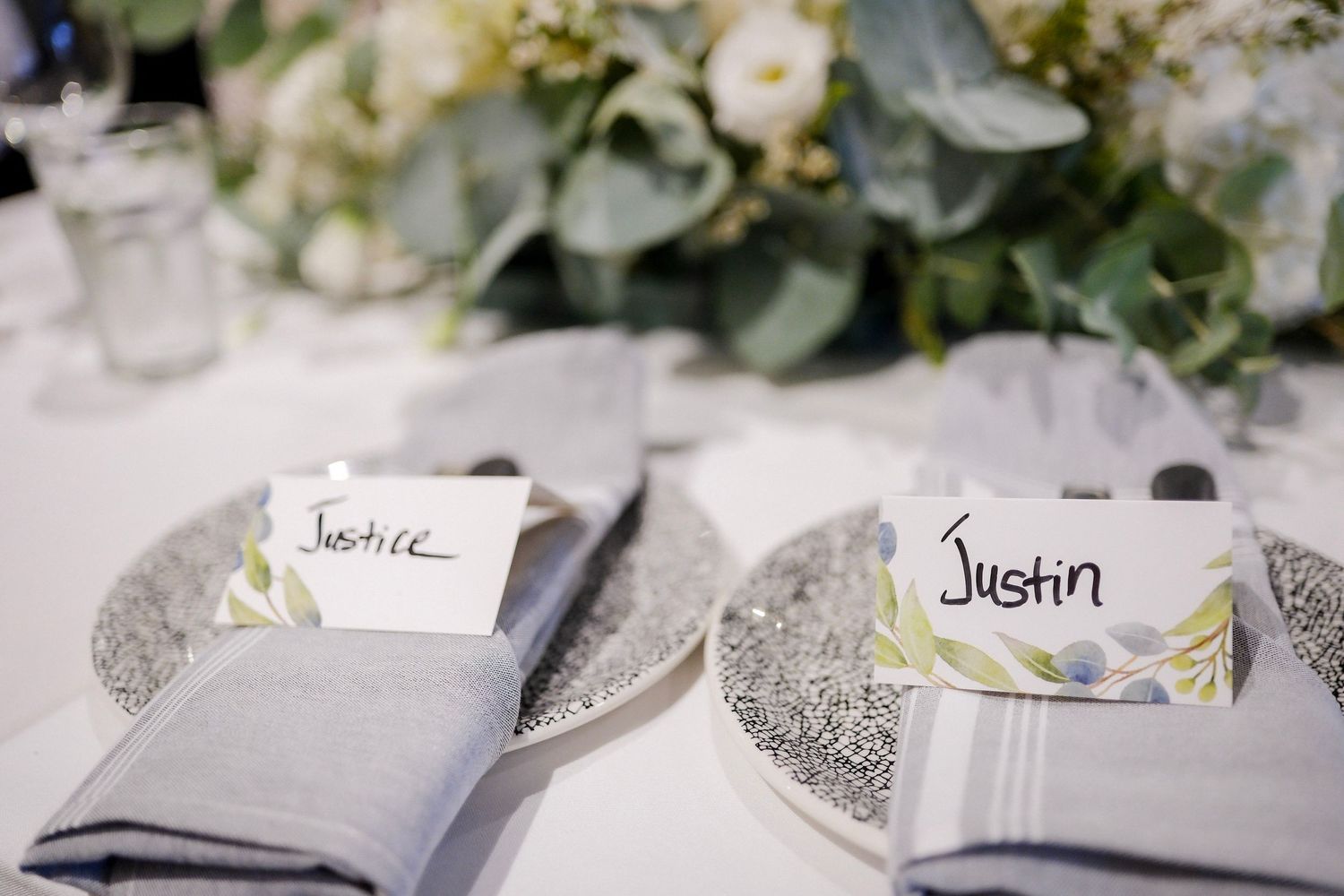Wedding place settings with name cards and silver napkins displayed beside white floral arrangements at a reception table.
