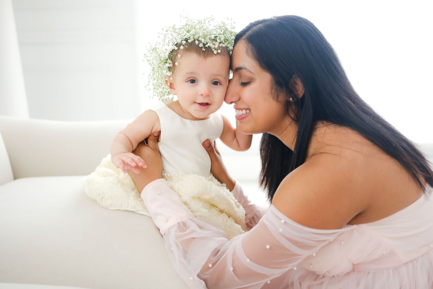 Mother and baby share sweet moment on white couch with baby wearing floral crown.