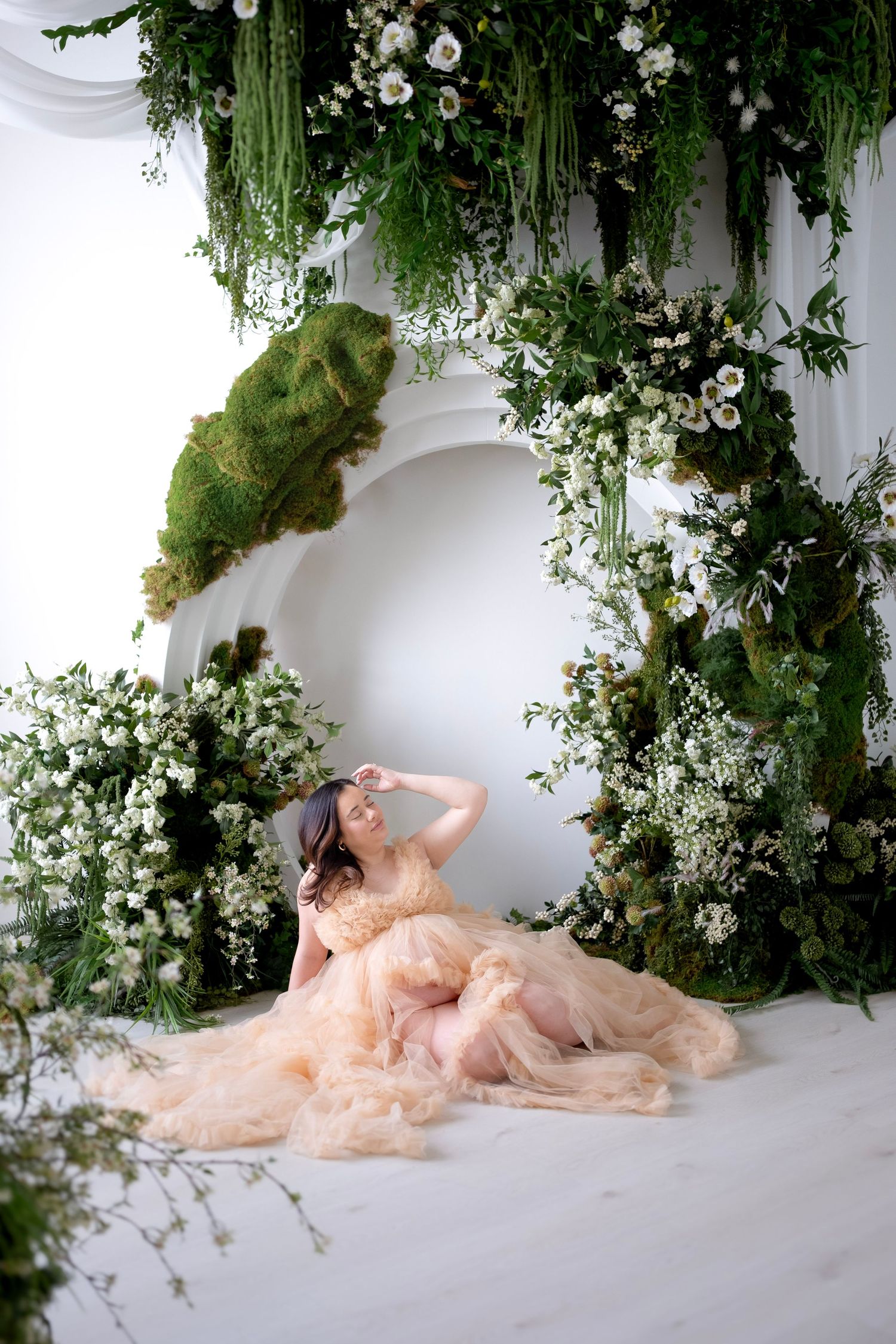 Woman in peach tulle gown poses among lush white flowers and green foliage in an elegant floral studio setting.