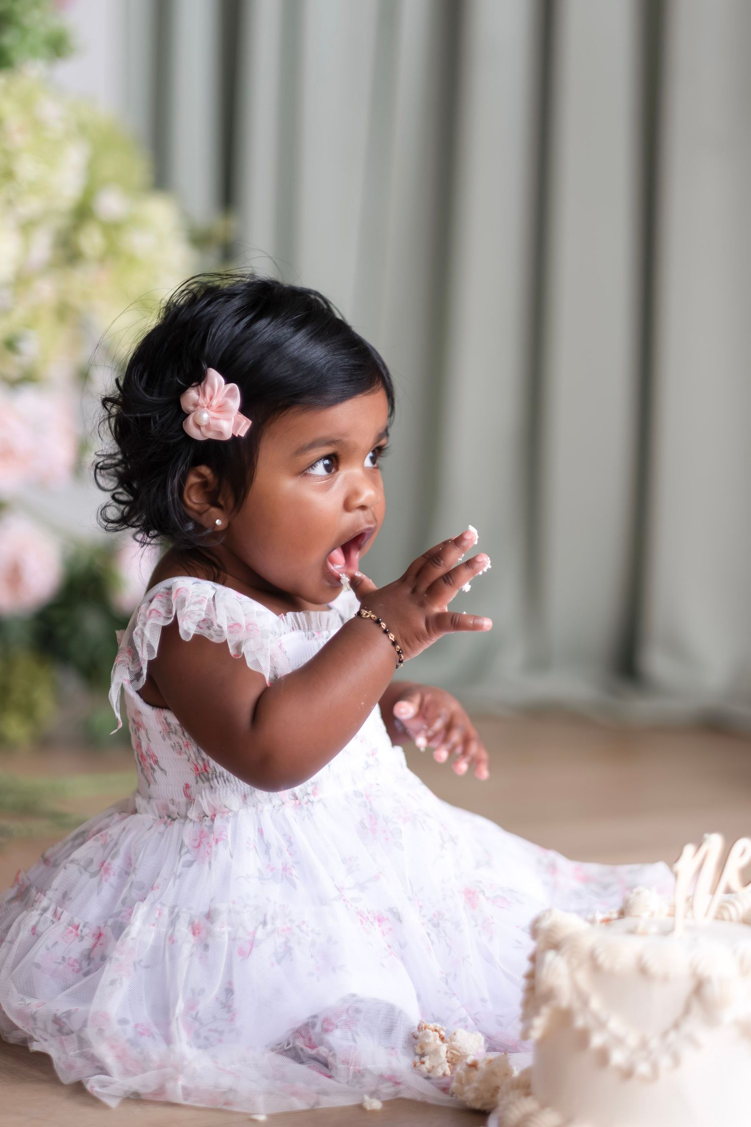 Toddler girl in white floral dress with pink hair bow enjoys her first birthday cake smash.