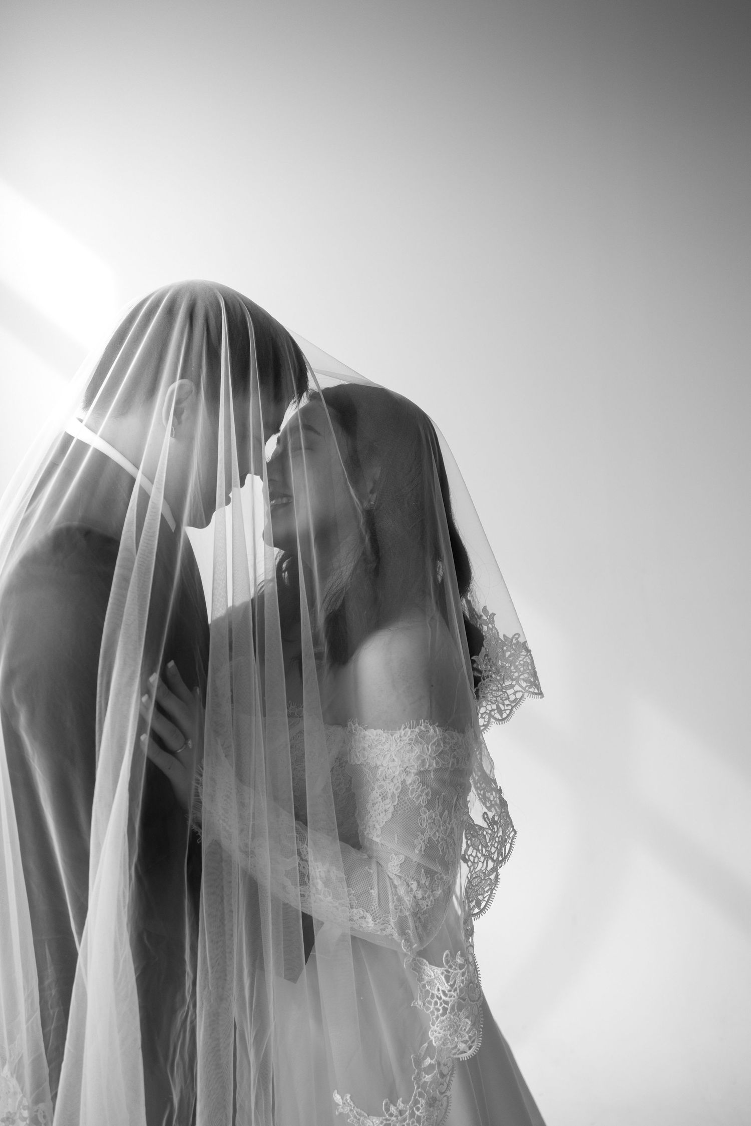 Black and white photo of bride and groom sharing a kiss under a flowing lace wedding veil in soft light.