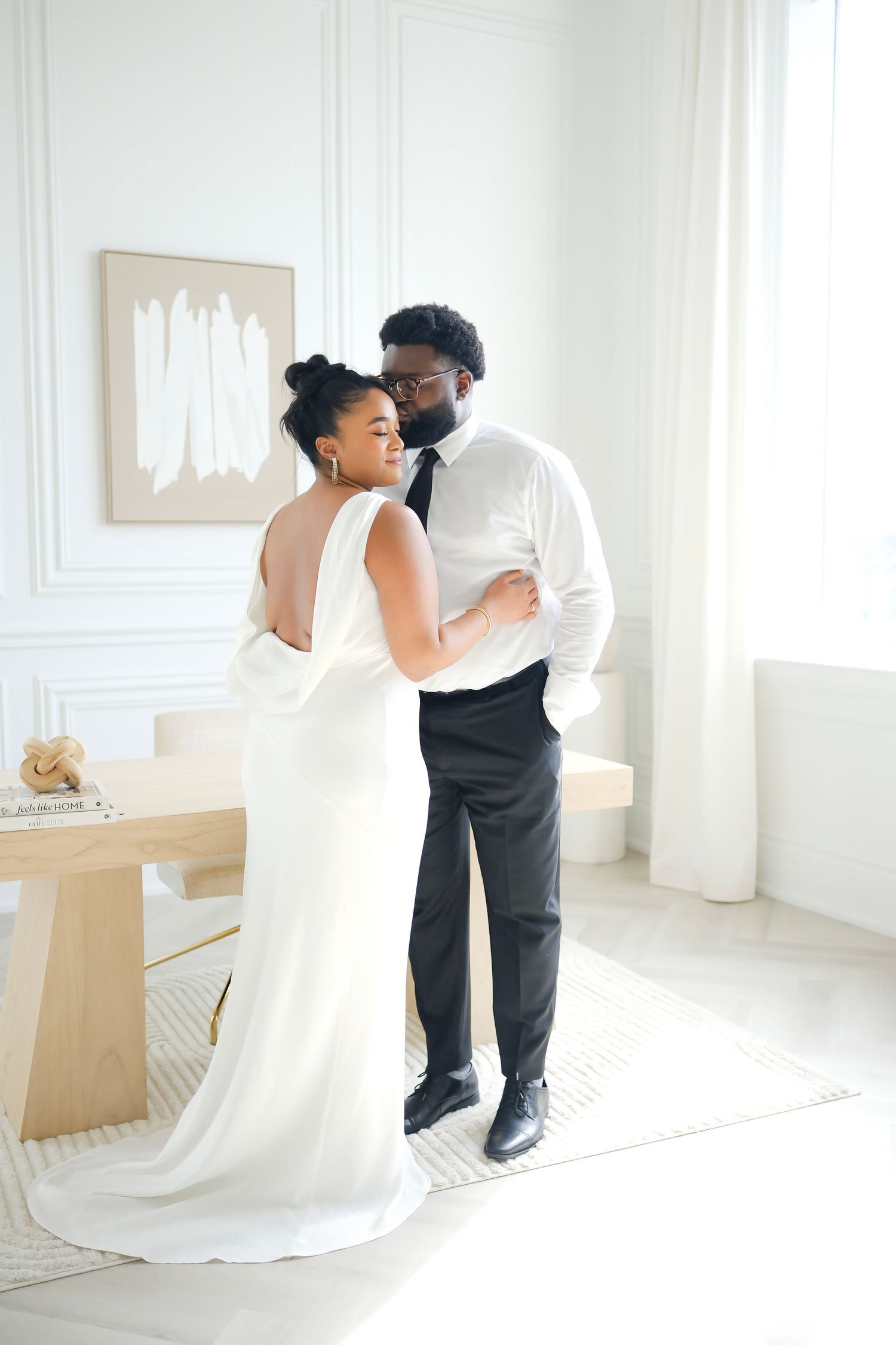 A couple shares an intimate moment in a bright white room, with the bride wearing a flowing white gown.
