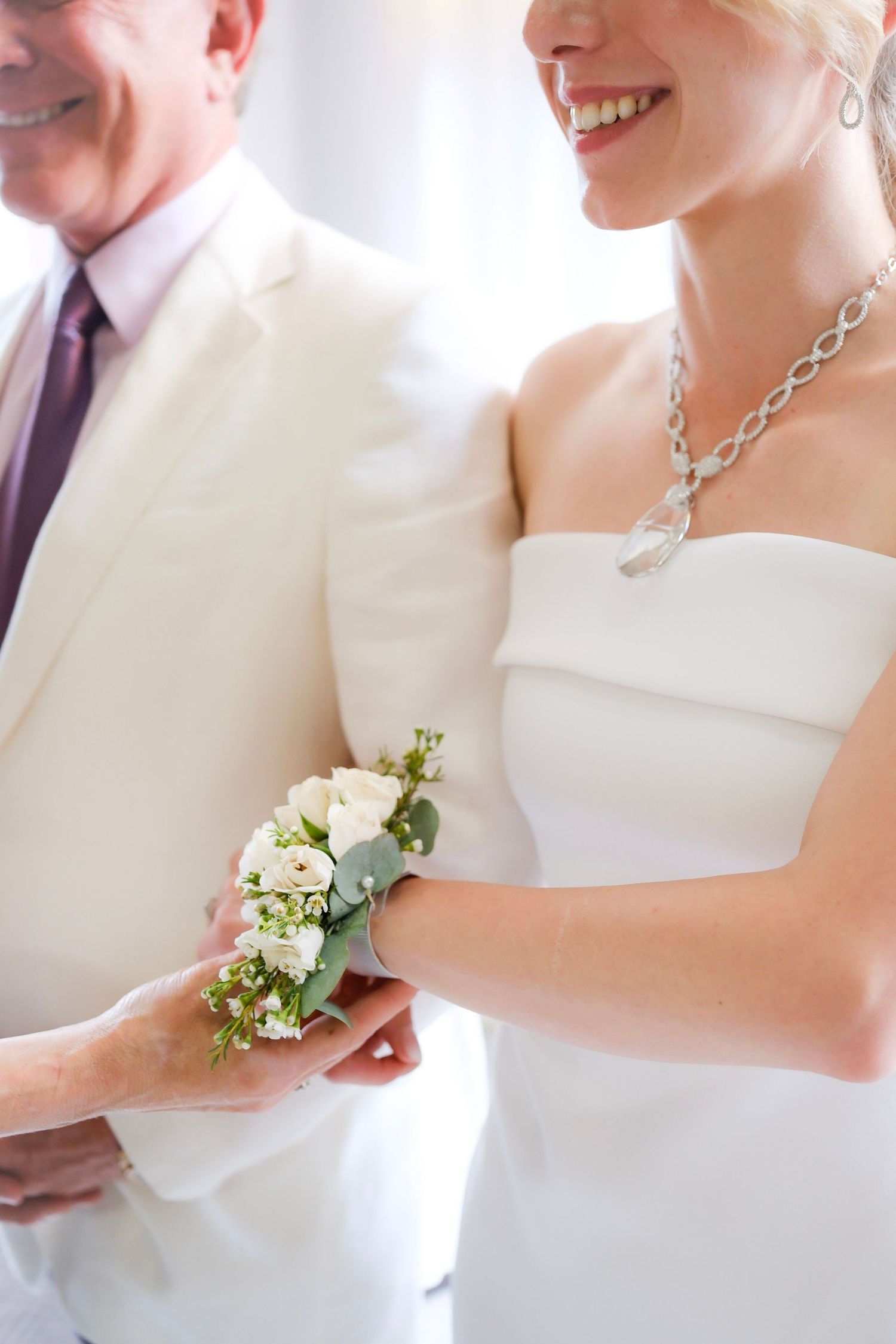A bride in a white dress wears a silver necklace and holds a delicate corsage of white and light blue flowers.