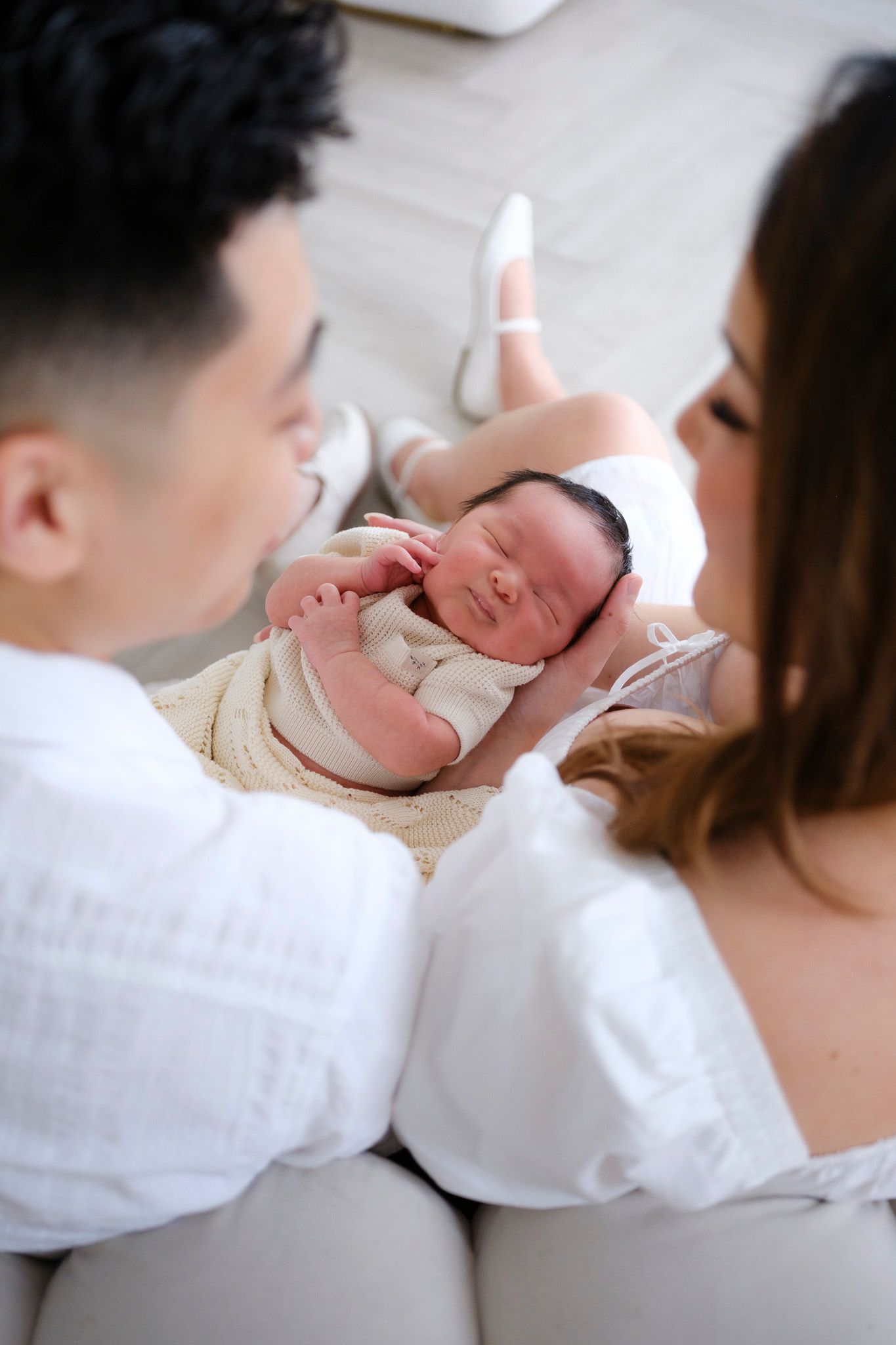 A newborn baby sleeps peacefully in white blankets while being cradled by parents in white clothing.