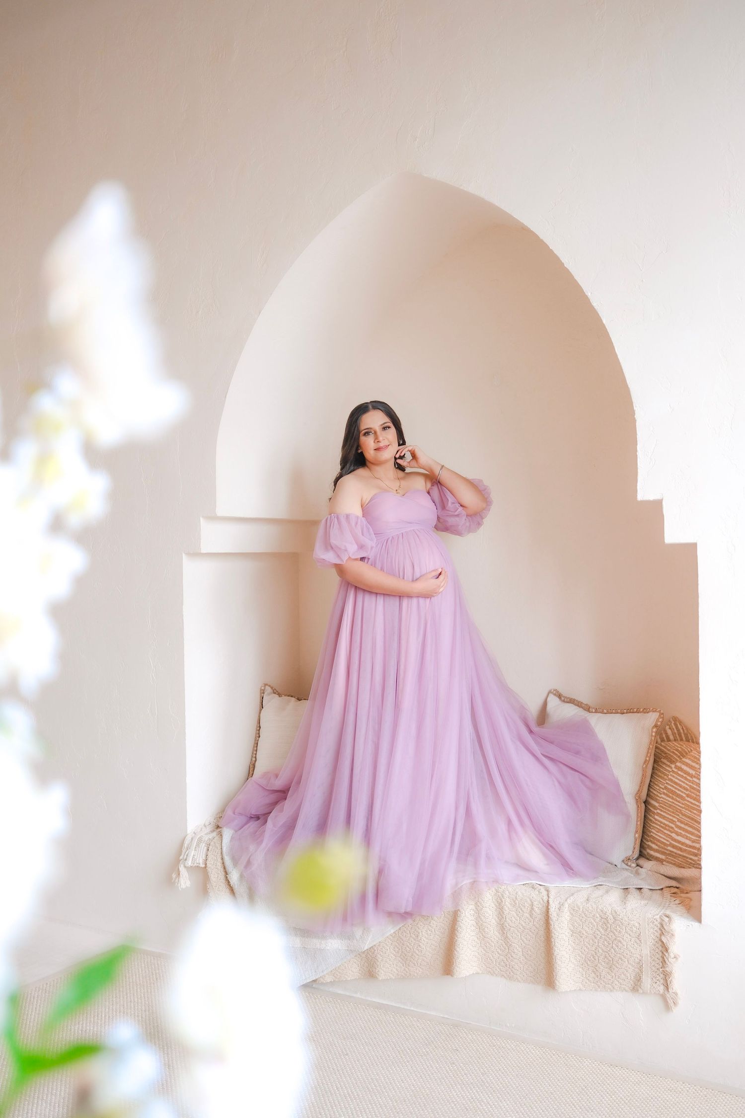 A flowing lavender off-shoulder maternity gown dress photographed in an elegant arched white alcove setting.