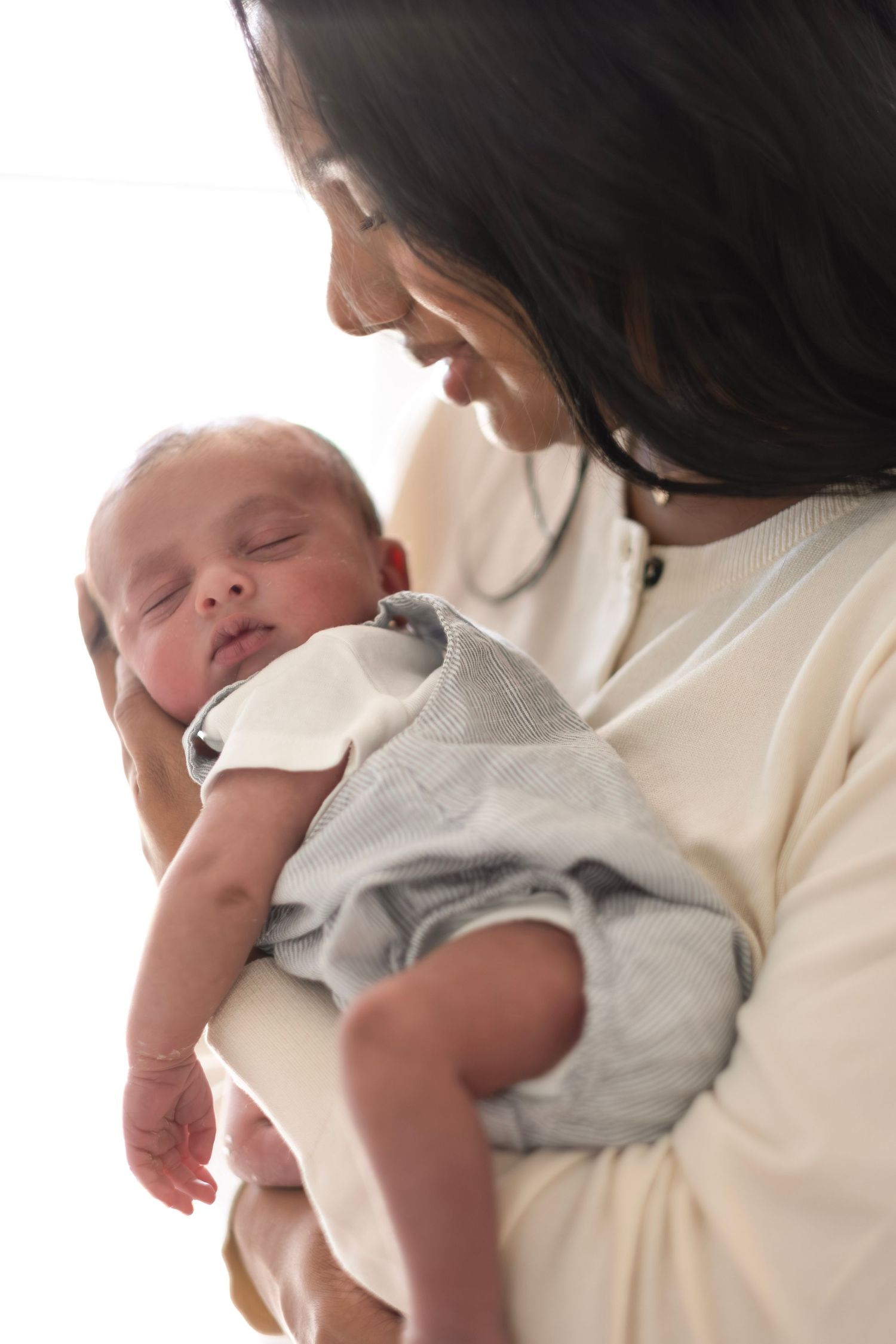 A mother tenderly holds her sleeping newborn baby dressed in a gray outfit against a bright background.