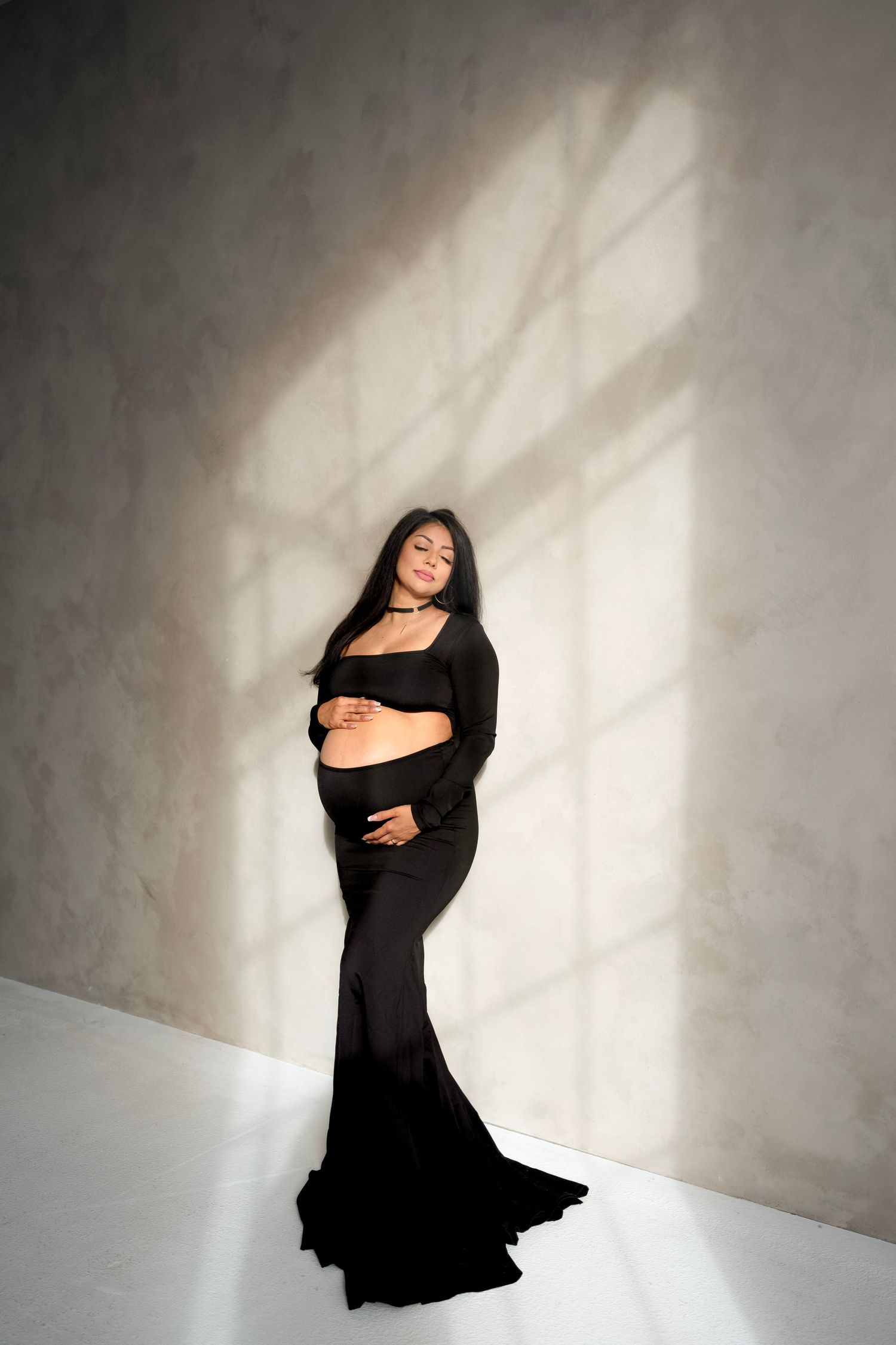 Artistic maternity photo featuring a woman in a flowing black dress against a white wall with dramatic shadows.