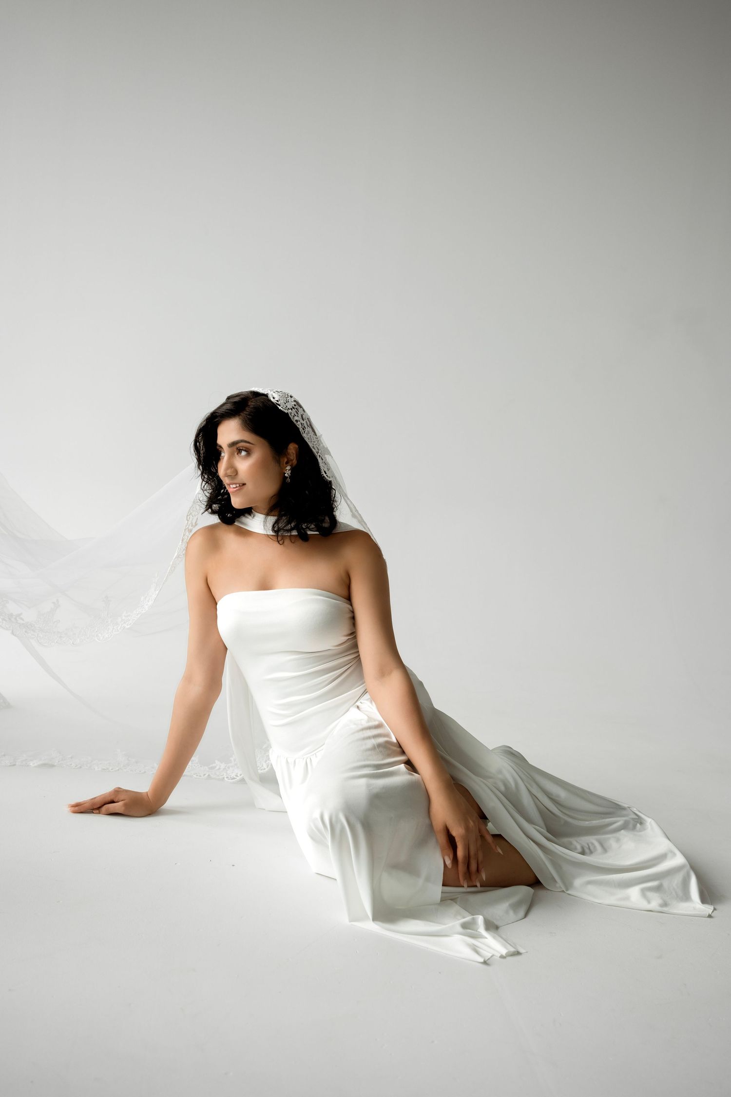 Elegant bride in strapless white satin gown and veil sitting gracefully on a white studio floor.