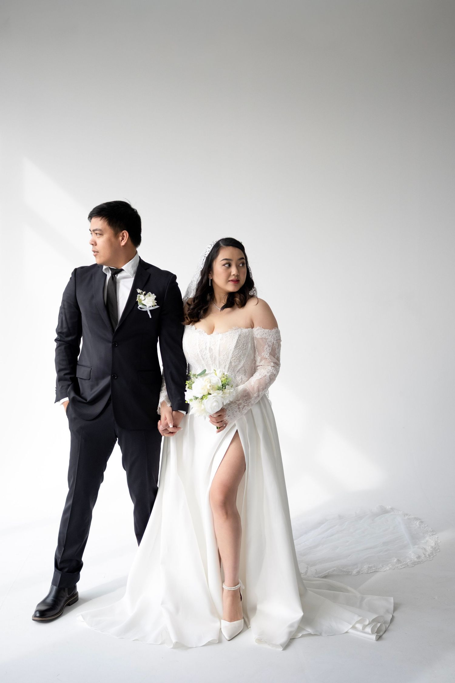 Asian couple in wedding attire, groom in black suit and bride in white off-shoulder gown holding bouquet.