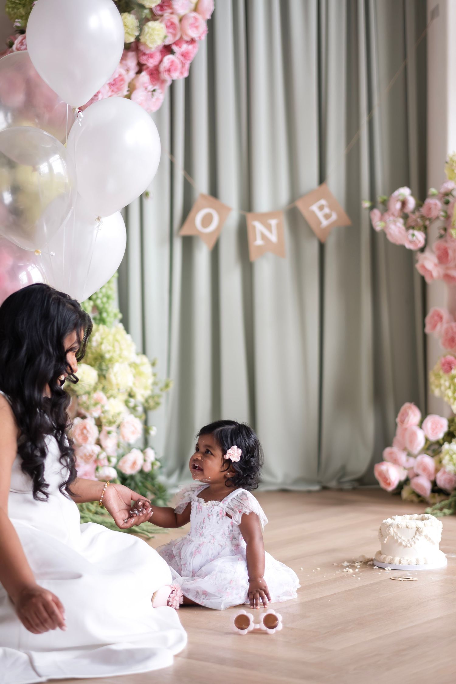 A mother sits with her baby girl near a first birthday cake under an ‘ONE’ banner with pink floral decor.