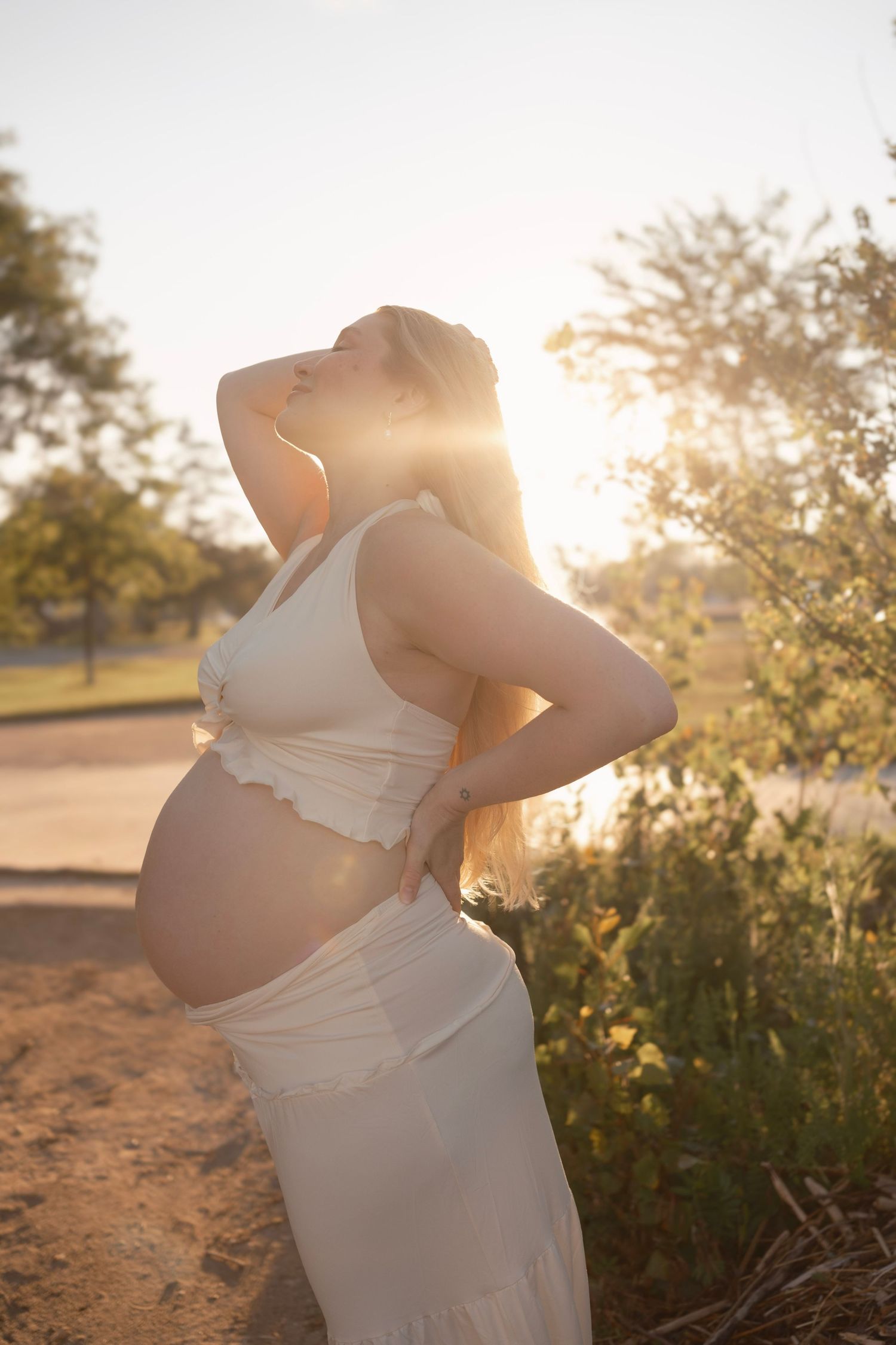 Pregnant woman in white crop top and skirt holding baby bump outdoors in golden sunset light.