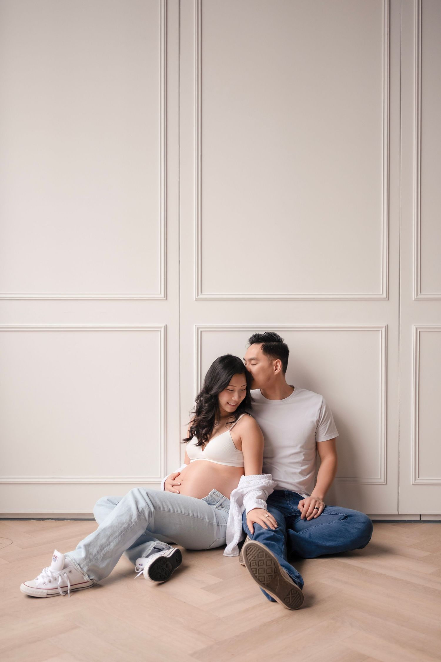 Pregnant Asian couple sitting on floor, man kissing woman's cheek, both in casual jeans and white tops.