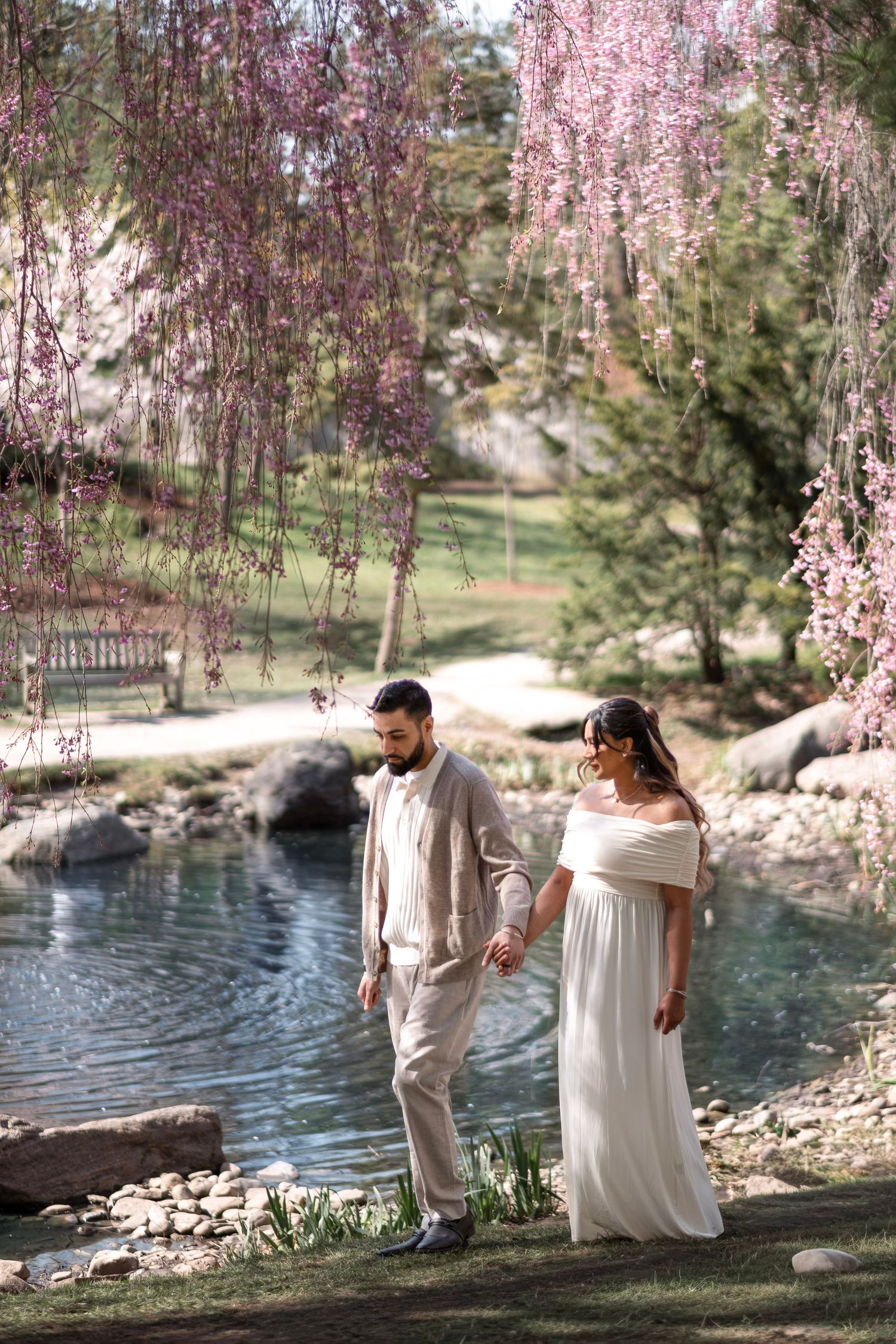 Couple holding hands by a serene pond under blooming pink weeping cherry trees, bride in white gown, groom in beige suit.