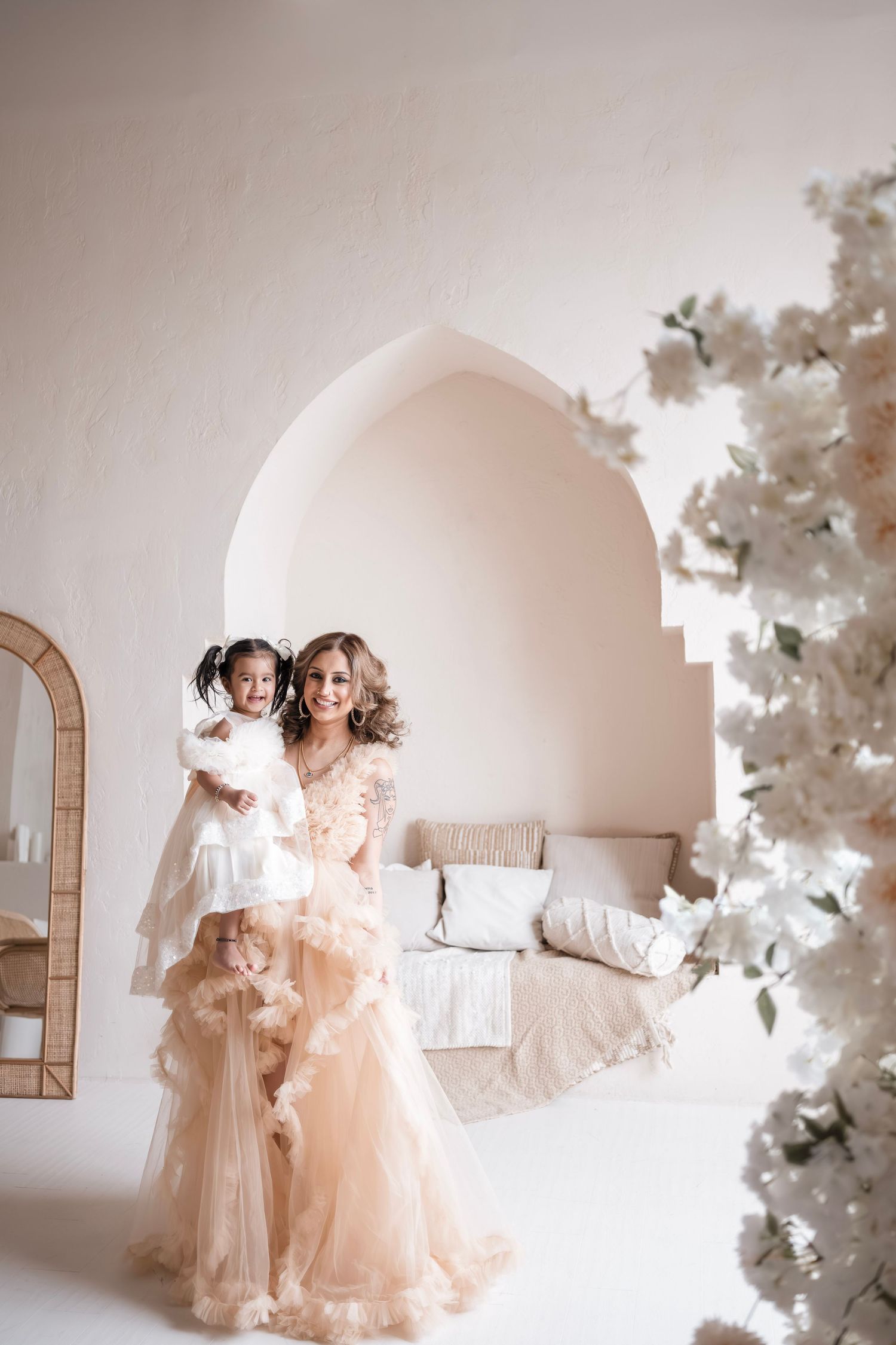 Woman in champagne tulle gown holds young girl in white dress, posing in elegant white room with floral decor.