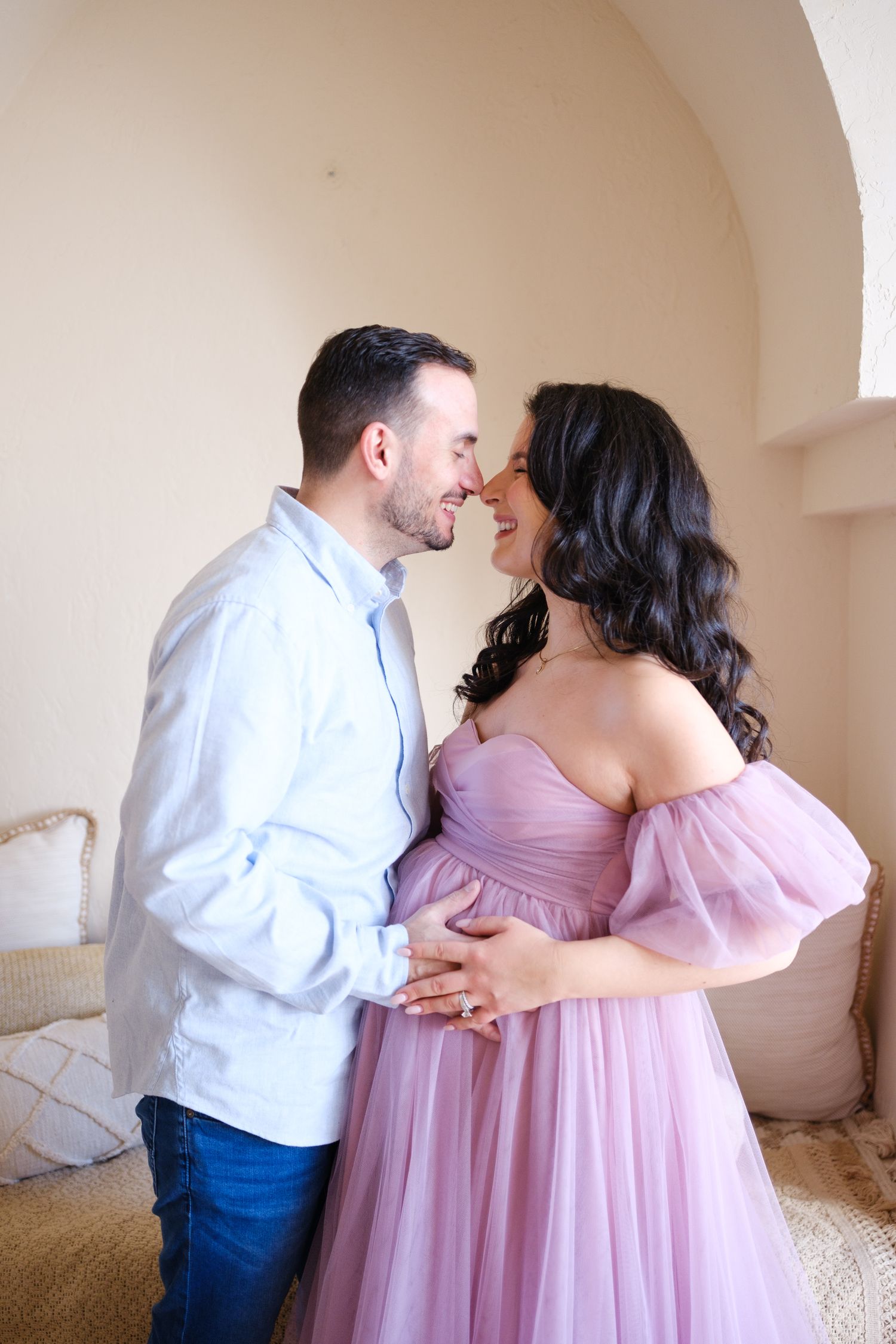 A couple shares an intimate moment in their home as they celebrate a pregnancy in a flowing pink off-shoulder dress.