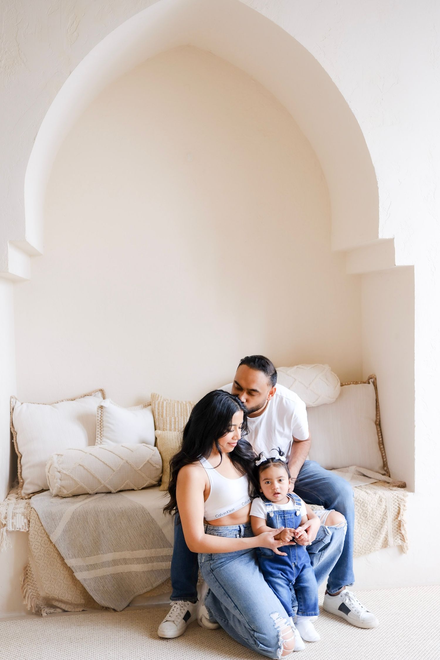 Family sits together on beige couch in minimalist white arched alcove with throw pillows.