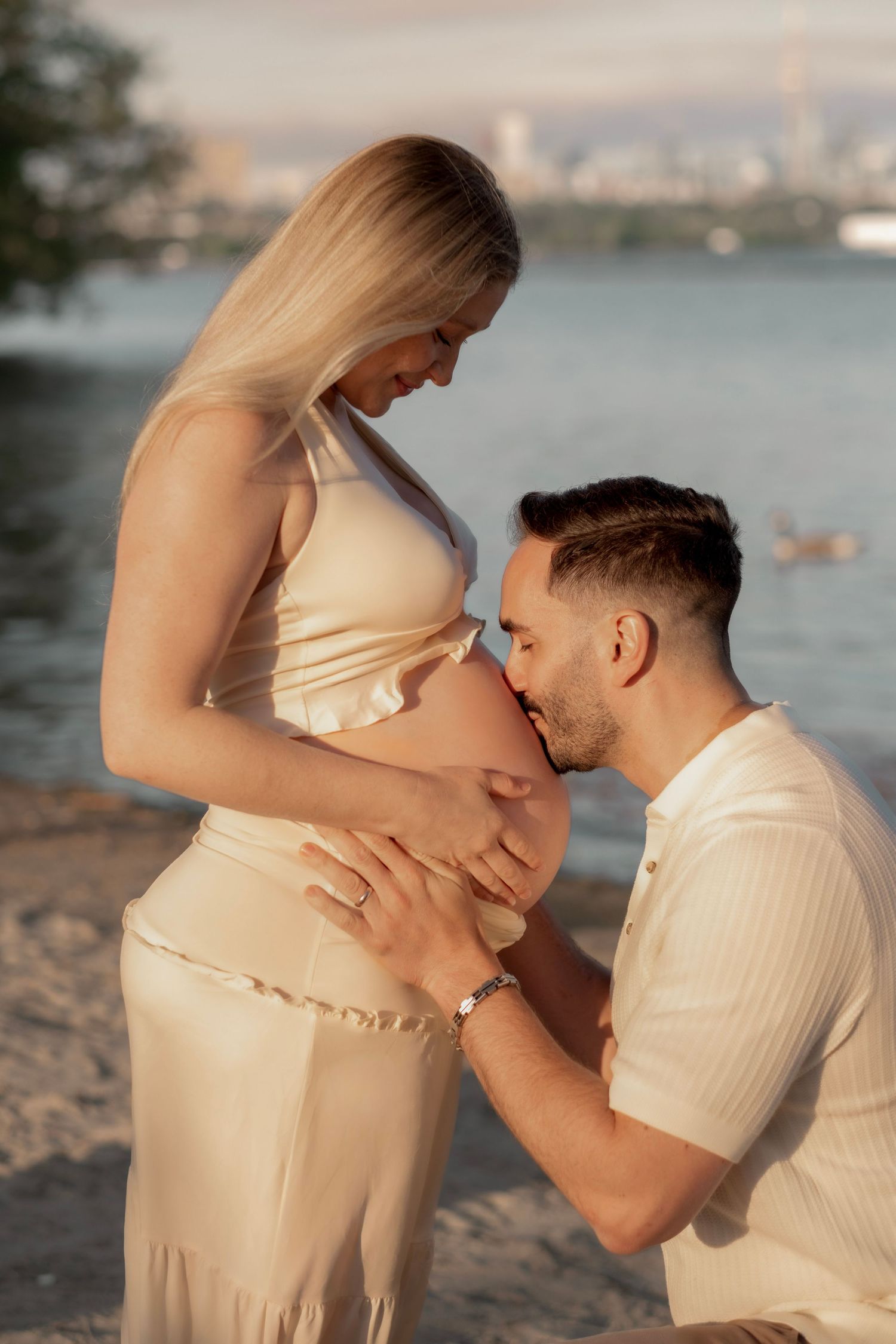 A man kneels on a beach kissing a pregnant woman's belly while she tenderly holds her bump near a waterfront.
