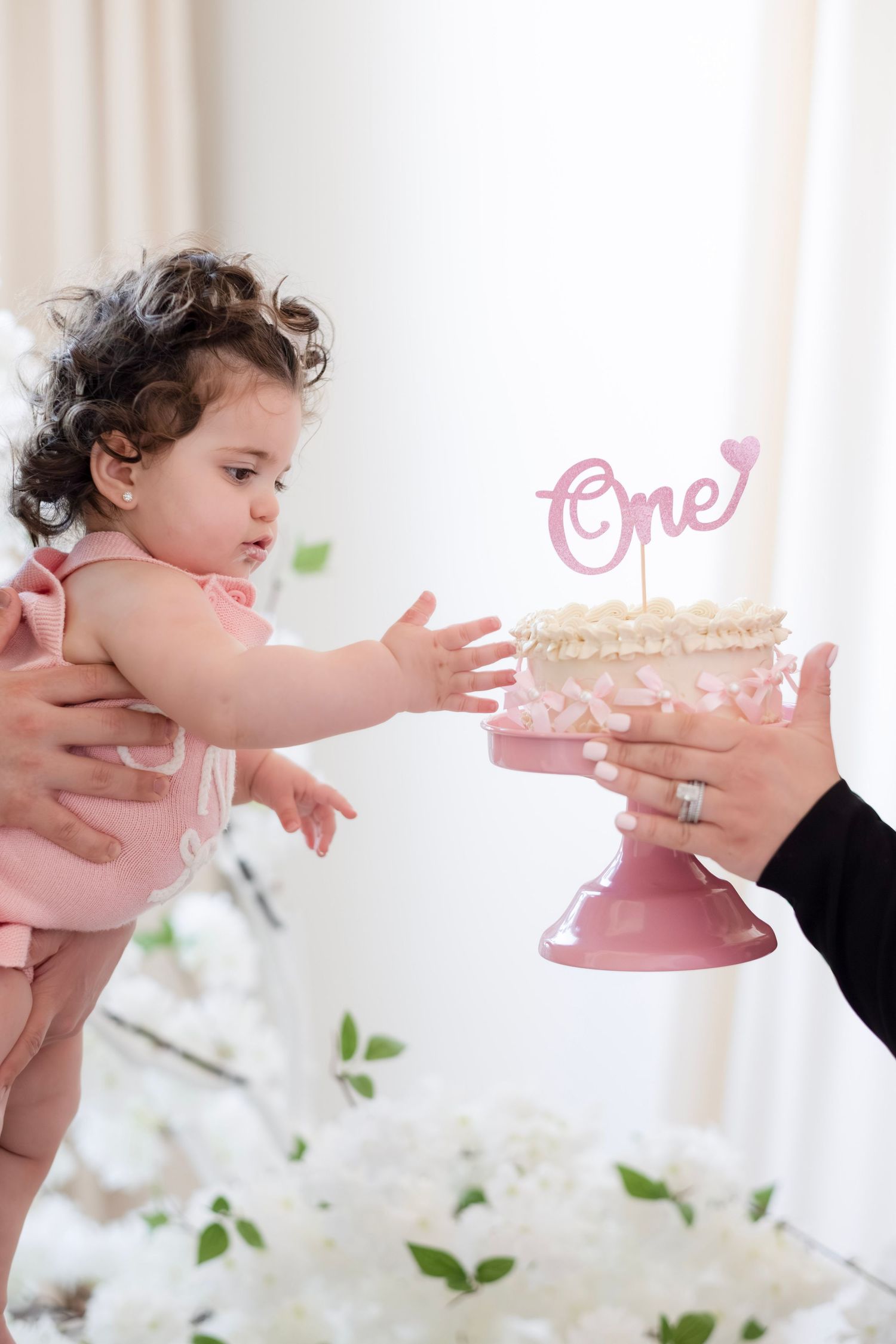 A baby girl in pink reaching for her first birthday cake with \