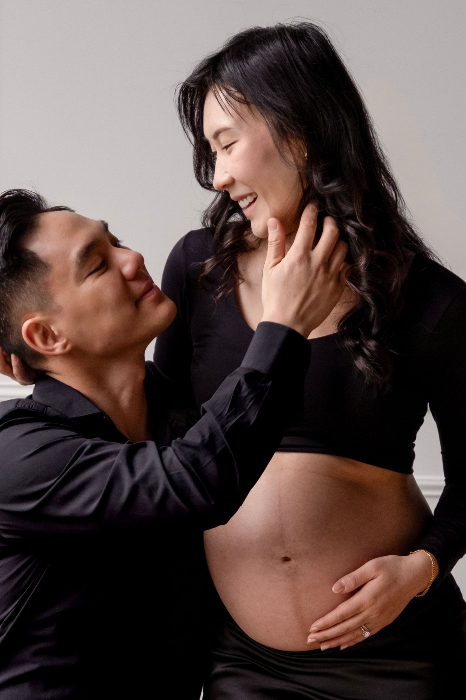 A couple in black outfits sharing a tender moment, with the woman visibly pregnant, smiling at each other in a studio.