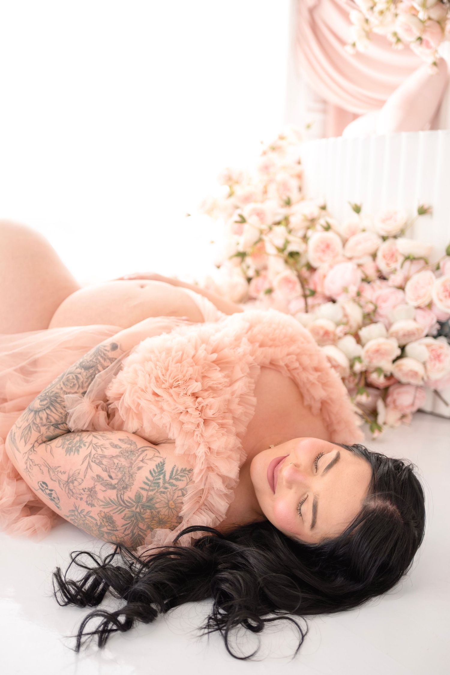 Pregnant woman with tattoos lying on white floor in pink robe surrounded by blush roses for maternity photo shoot.
