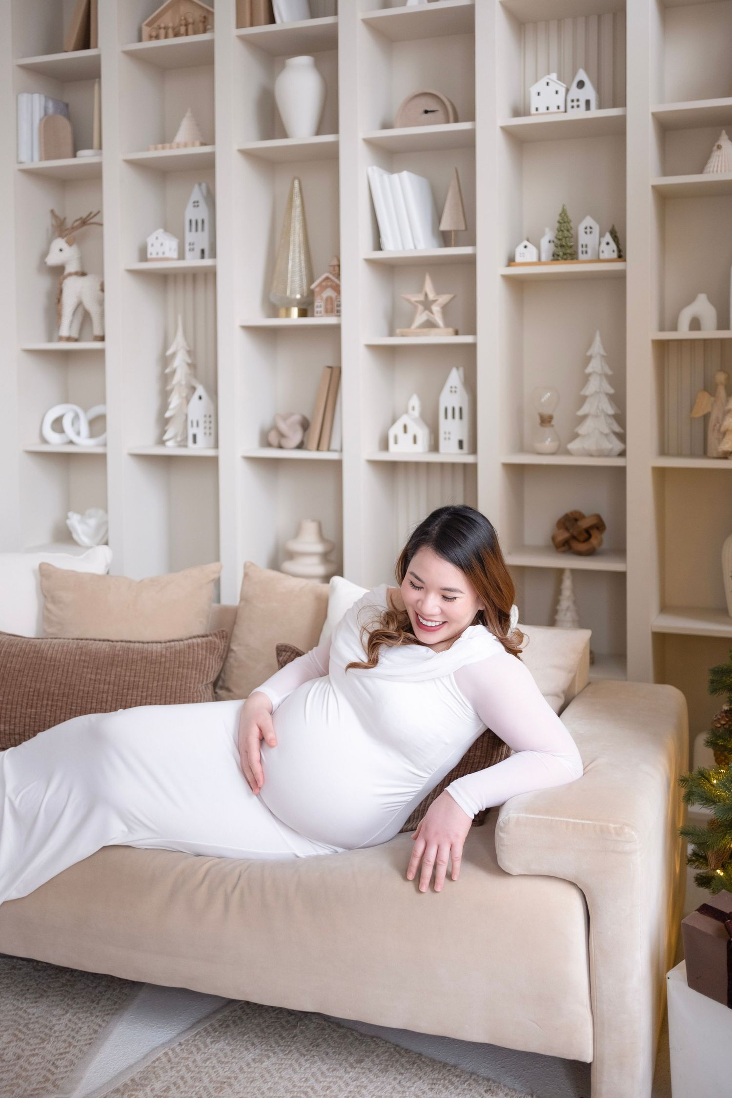 A person relaxes on a beige couch against a wall of white decorative shelves filled with miniature houses and ornaments.