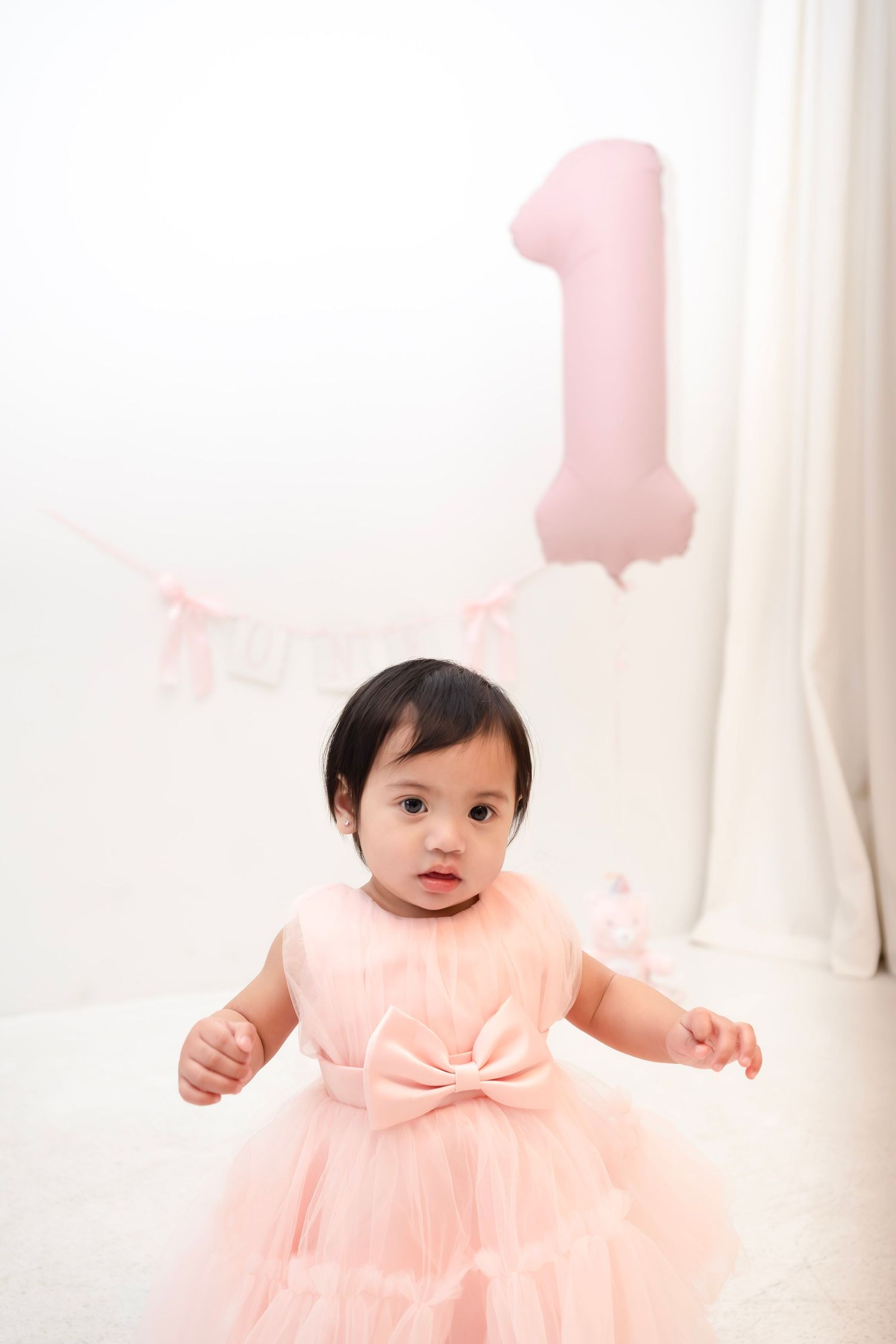 A baby girl in a pink tulle dress celebrates her first birthday with a number one balloon and bunting backdrop.