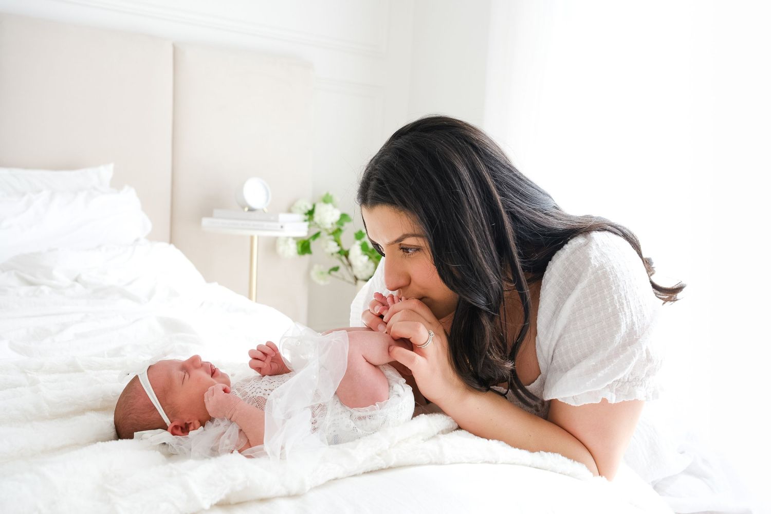 A mother lies on white bedding while gazing lovingly at her newborn baby in a bright, serene bedroom setting.