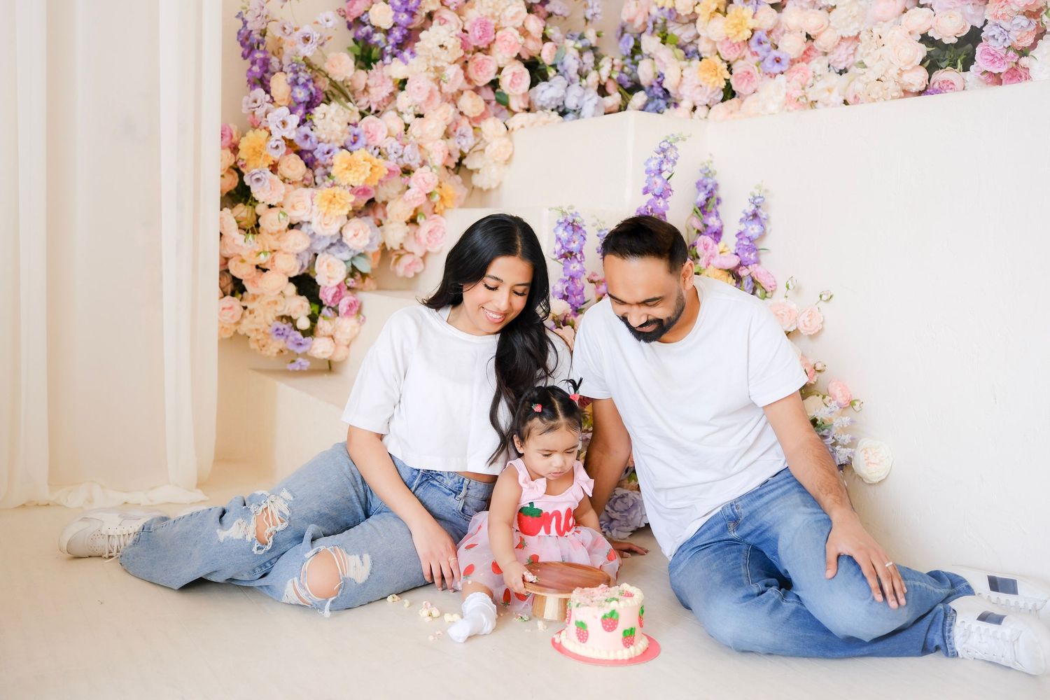 Family in matching white tops and jeans sitting on floor with birthday cake surrounded by pink and purple floral backdrop.