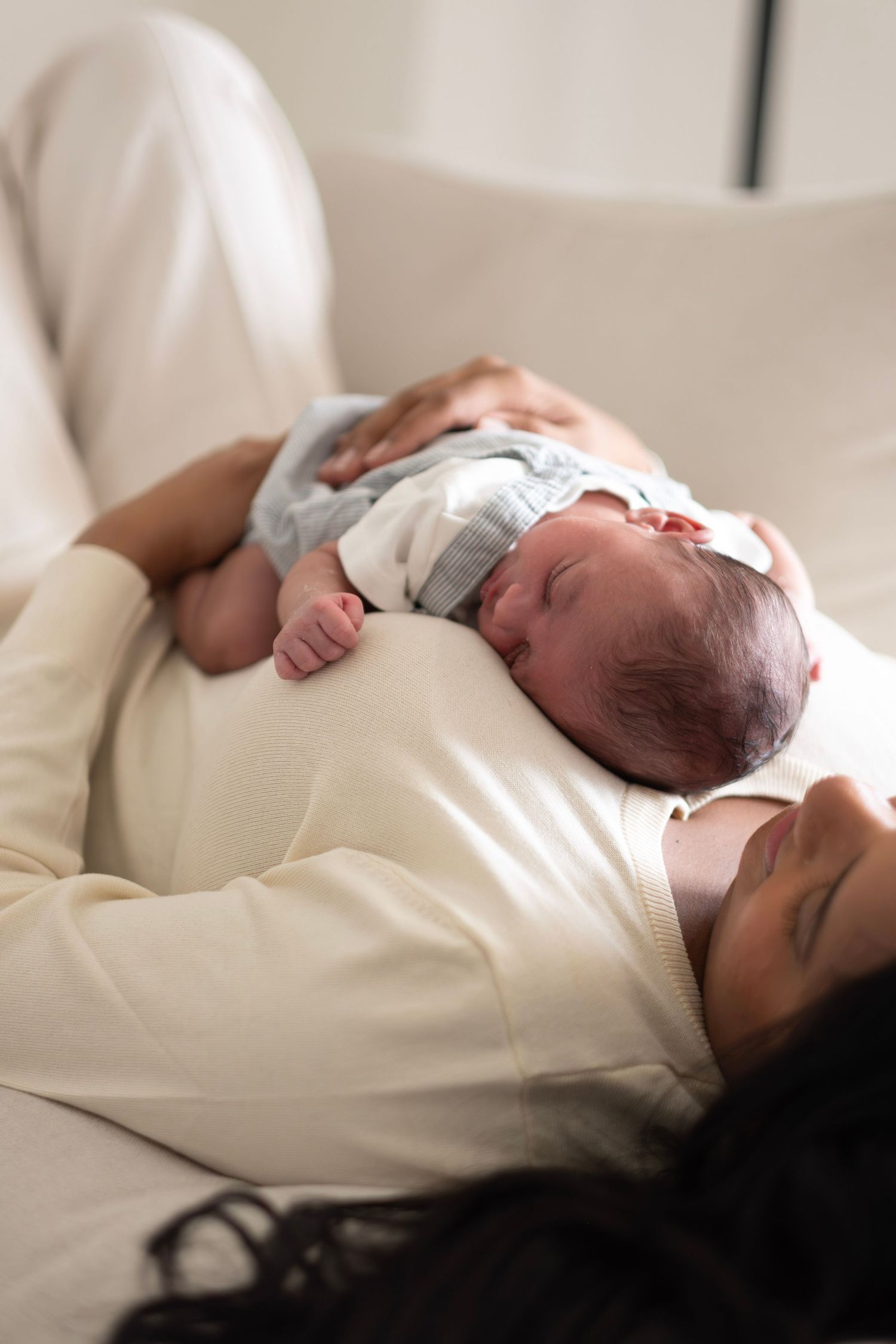 Mother resting on a cream sofa while cradling her sleeping newborn baby dressed in a white and blue outfit.