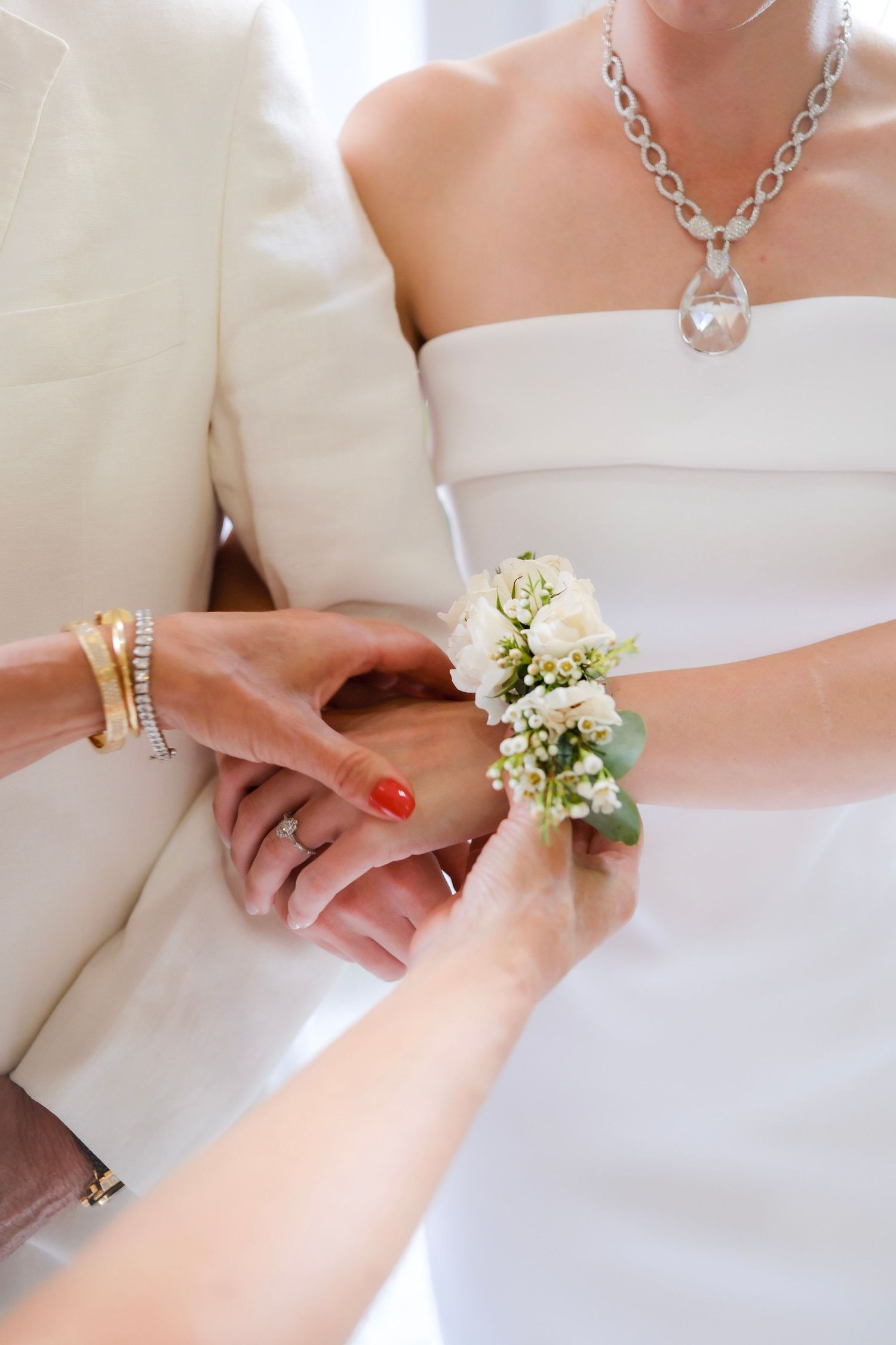 Someone helps attach a delicate white floral corsage to a wrist while wearing elegant wedding attire and jewelry.