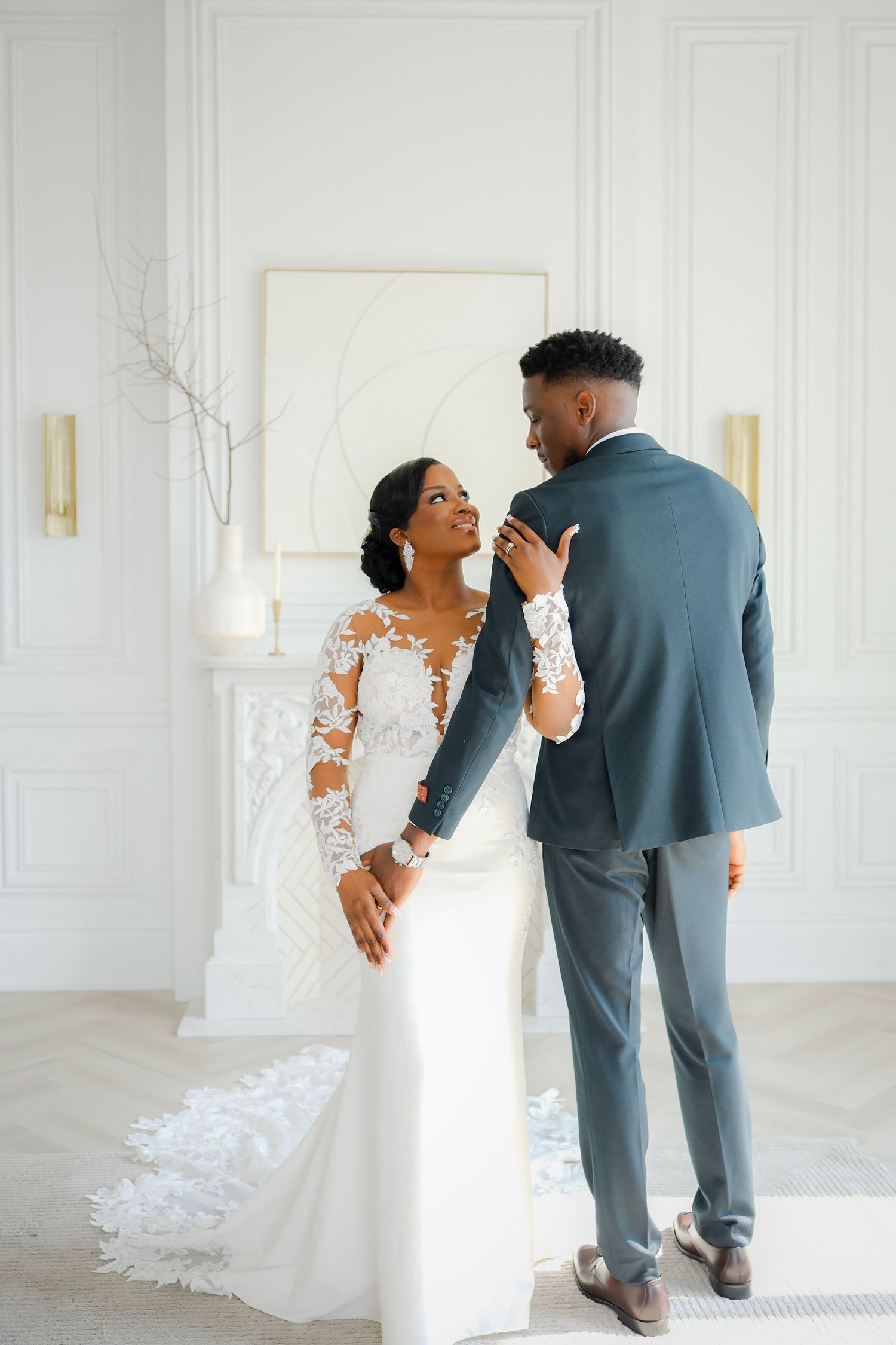 An elegant couple in wedding attire share a tender moment in a bright white room with minimalist decor.