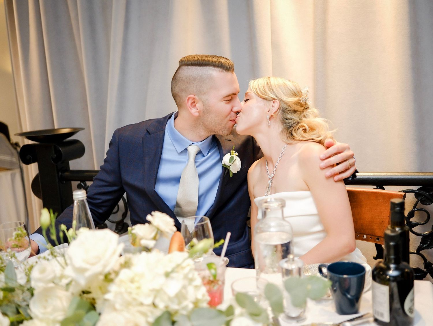 A bride and groom share a romantic kiss at their wedding reception with elegant floral centerpieces on the table.