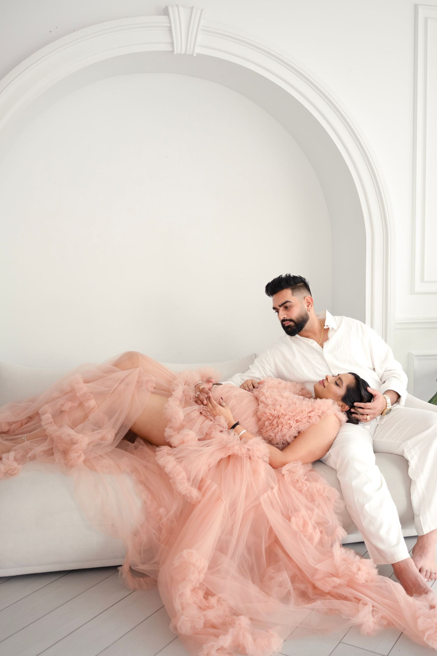 A couple in formal attire poses romantically in a white room with the woman wearing a flowing peach tulle gown.