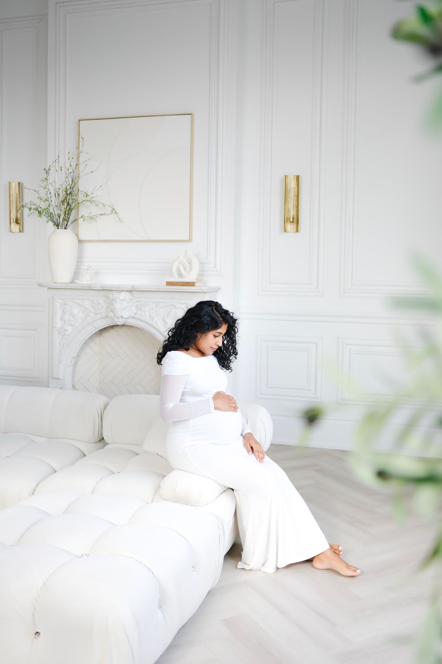 Expectant mother in white dress sits peacefully on elegant white couch in bright minimalist room with marble fireplace.