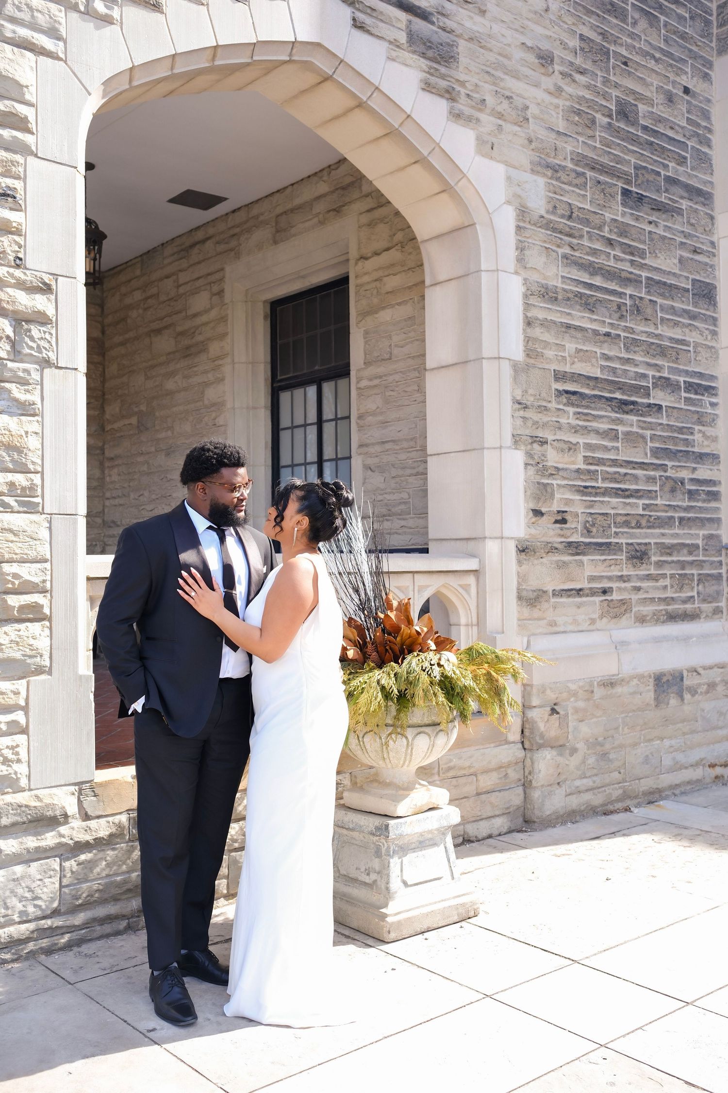 A couple in wedding attire share an intimate moment under a stone archway decorated with a flower planter.