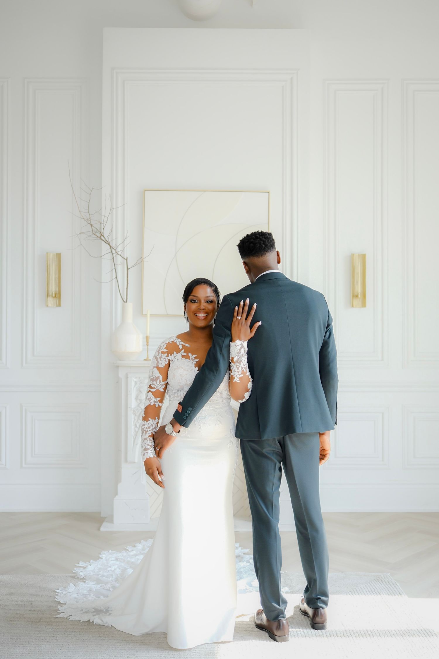 A beautiful bride in a white lace sleeve gown showing her ring while standing with her groom in a minimalist white room.