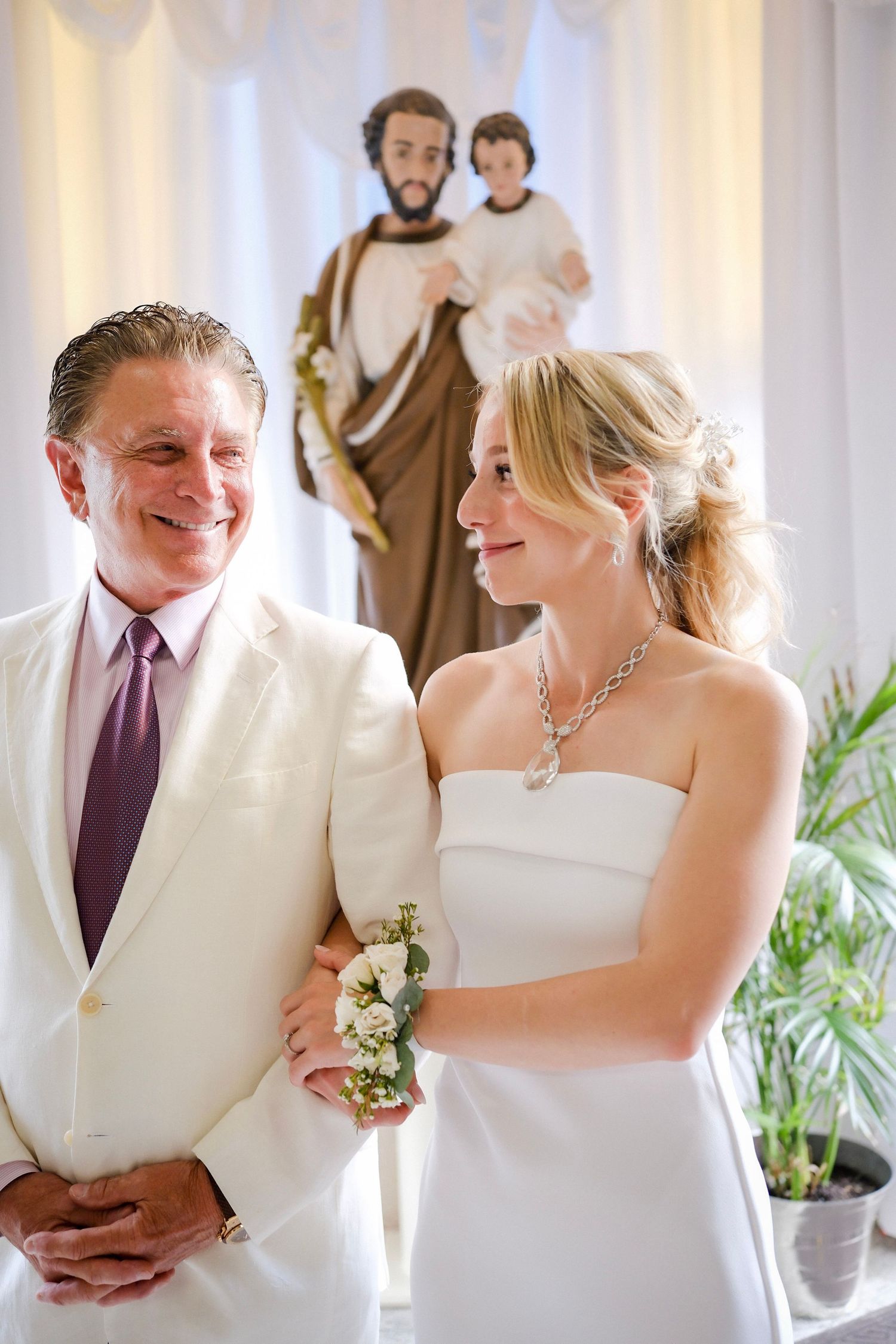 A couple in formal white attire share a joyful moment during their wedding ceremony with a religious statue behind them.