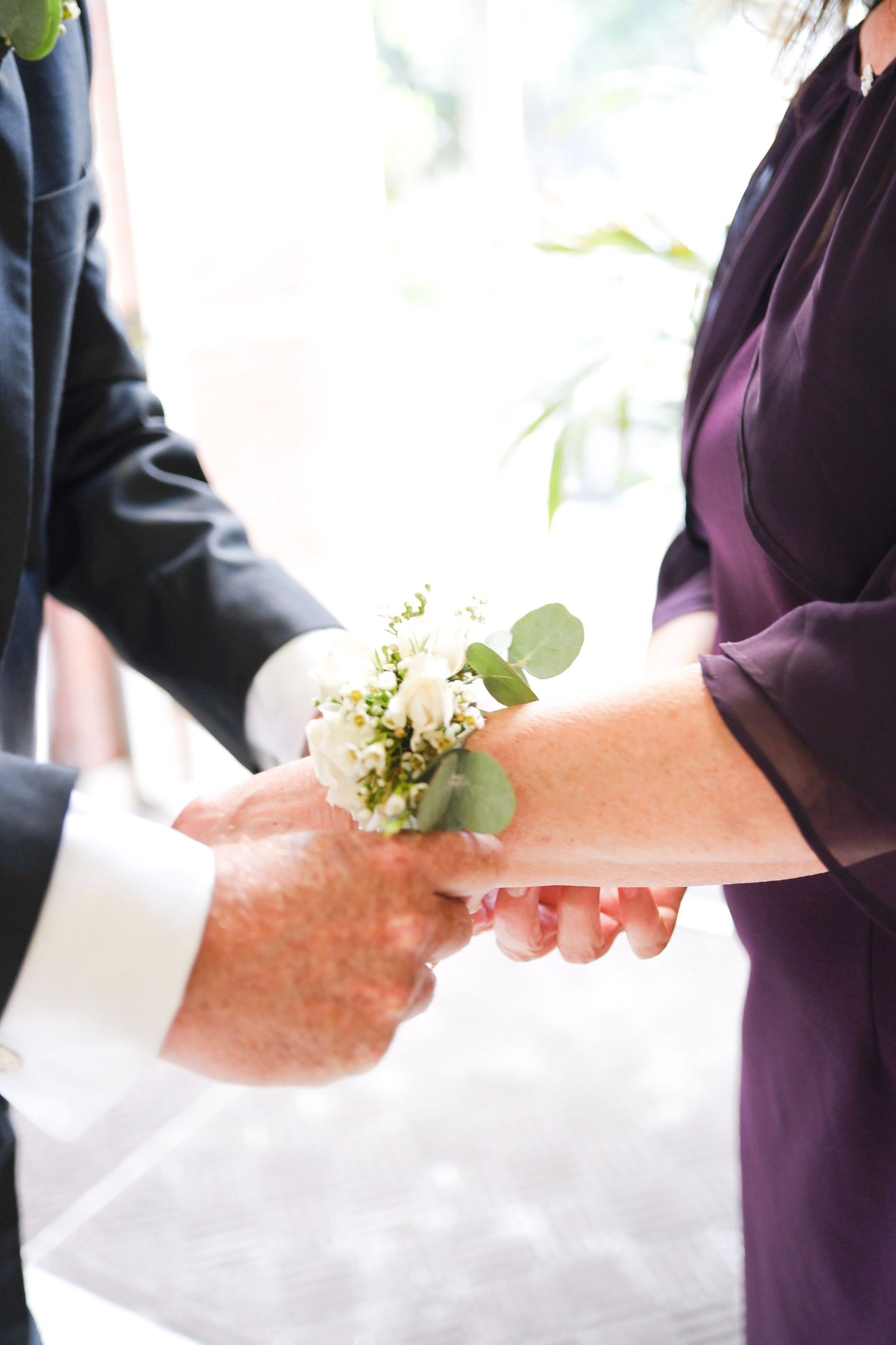 Two people hold hands during a wedding ceremony with a delicate white floral boutonniere visible between them.