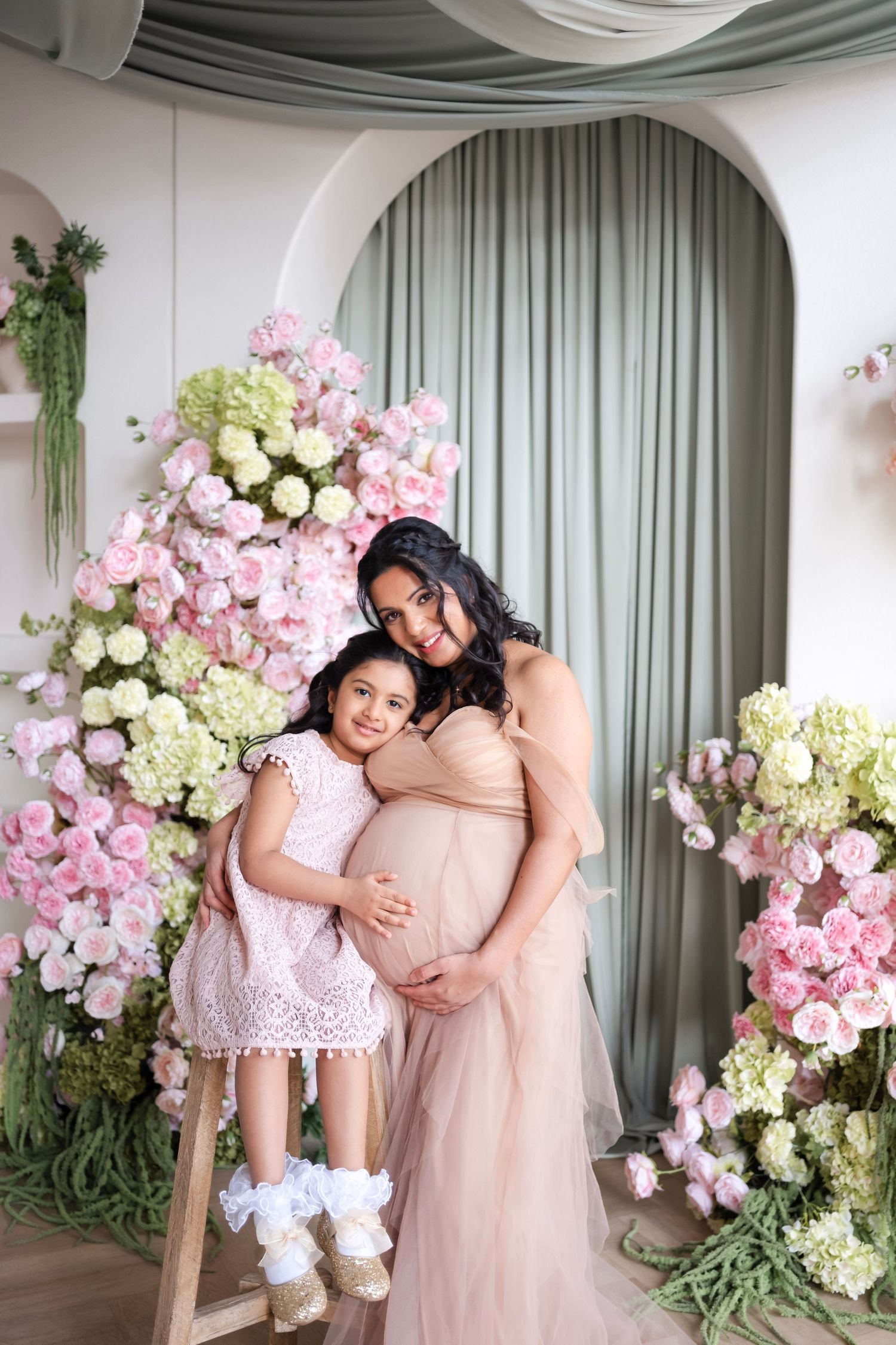 Pregnant woman in blush gown hugs young girl in pink dress, surrounded by floral backdrop of roses and hydrangeas.