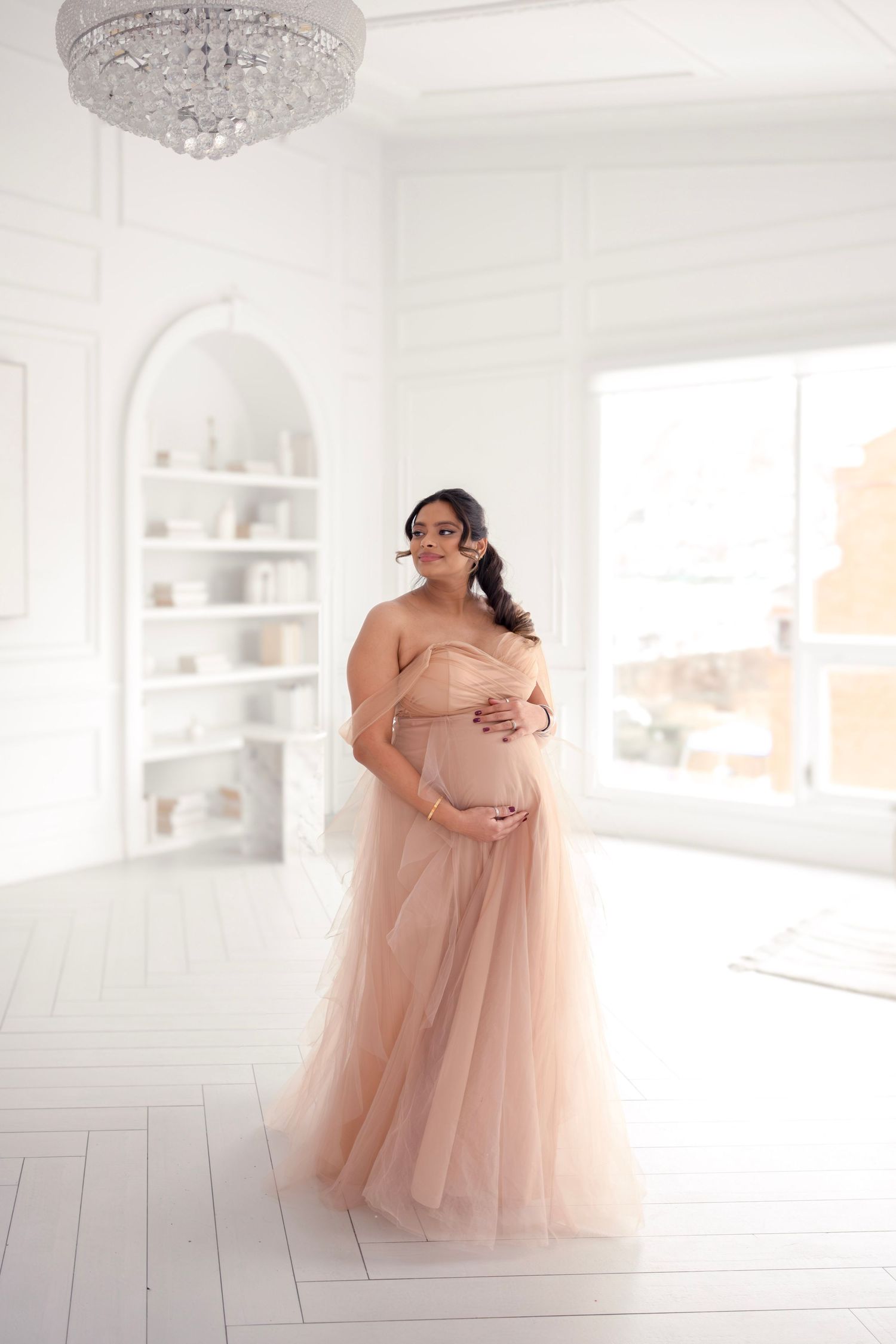 Romantic maternity photoshoot in white studio with flowing peach tulle gown and crystal chandelier.