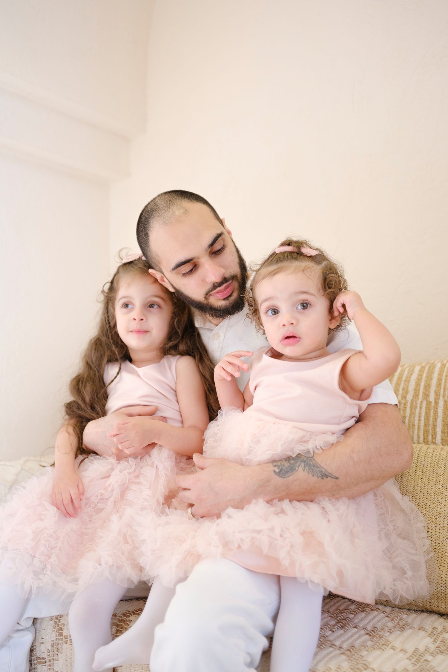 A tender family moment showing two young girls in pink tulle dresses sitting together on a cream-colored couch.
