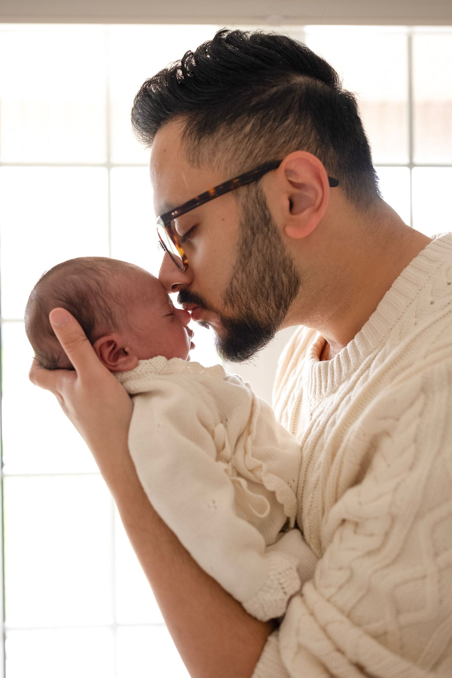 Bearded father with glasses gently kissing his newborn baby while holding them up near a bright window.