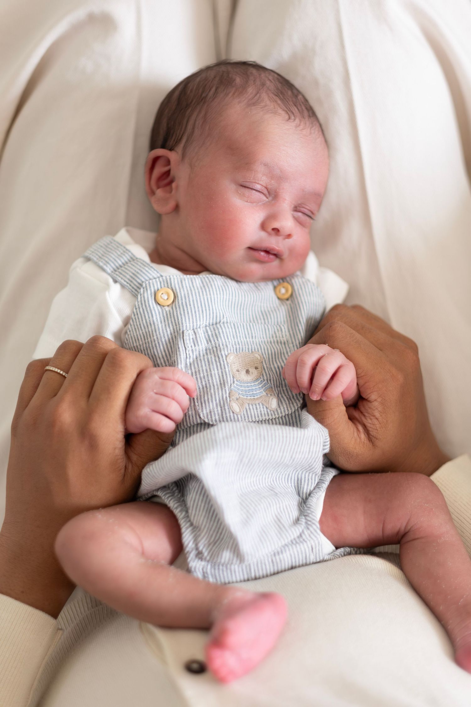 Newborn baby sleeping peacefully in parent's arms wearing a light blue striped romper with bear embroidery.