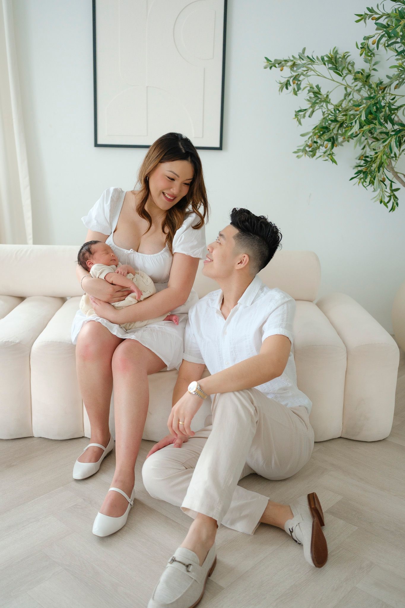 A family sitting together on a cream-colored couch in a bright minimalist living room with indoor plants.
