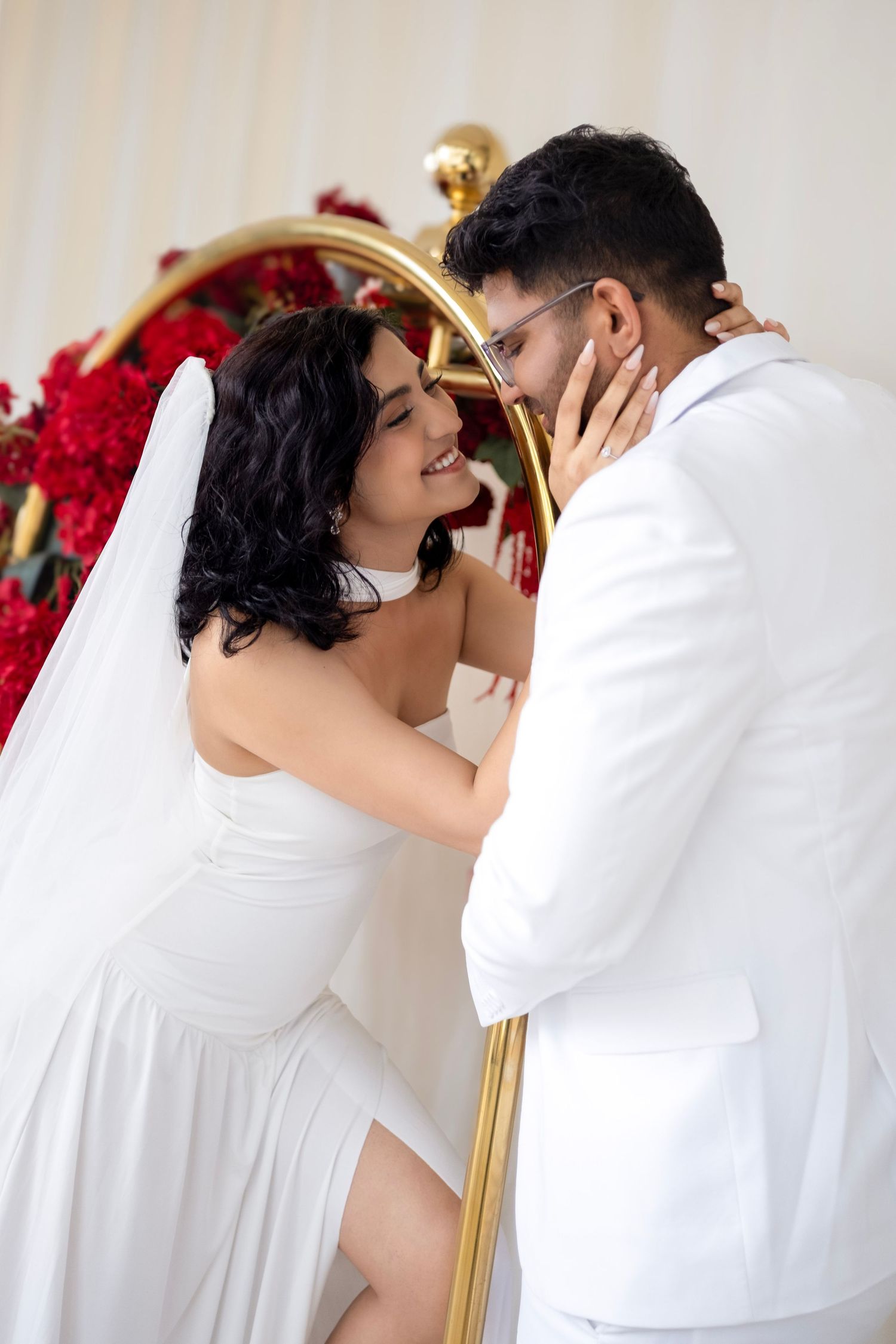 Bride and groom share a romantic moment, dressed in white wedding attire with red floral backdrop.