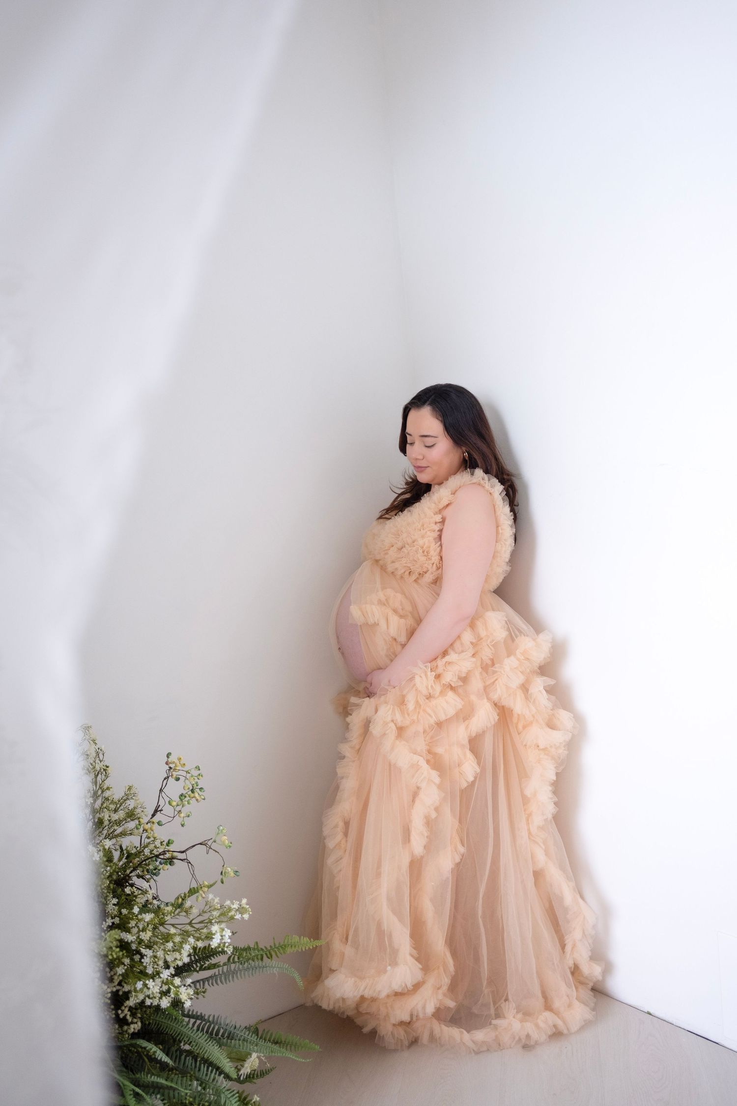 Pregnant woman in peach ruffled tulle maternity gown standing in white corner studio with flowers.