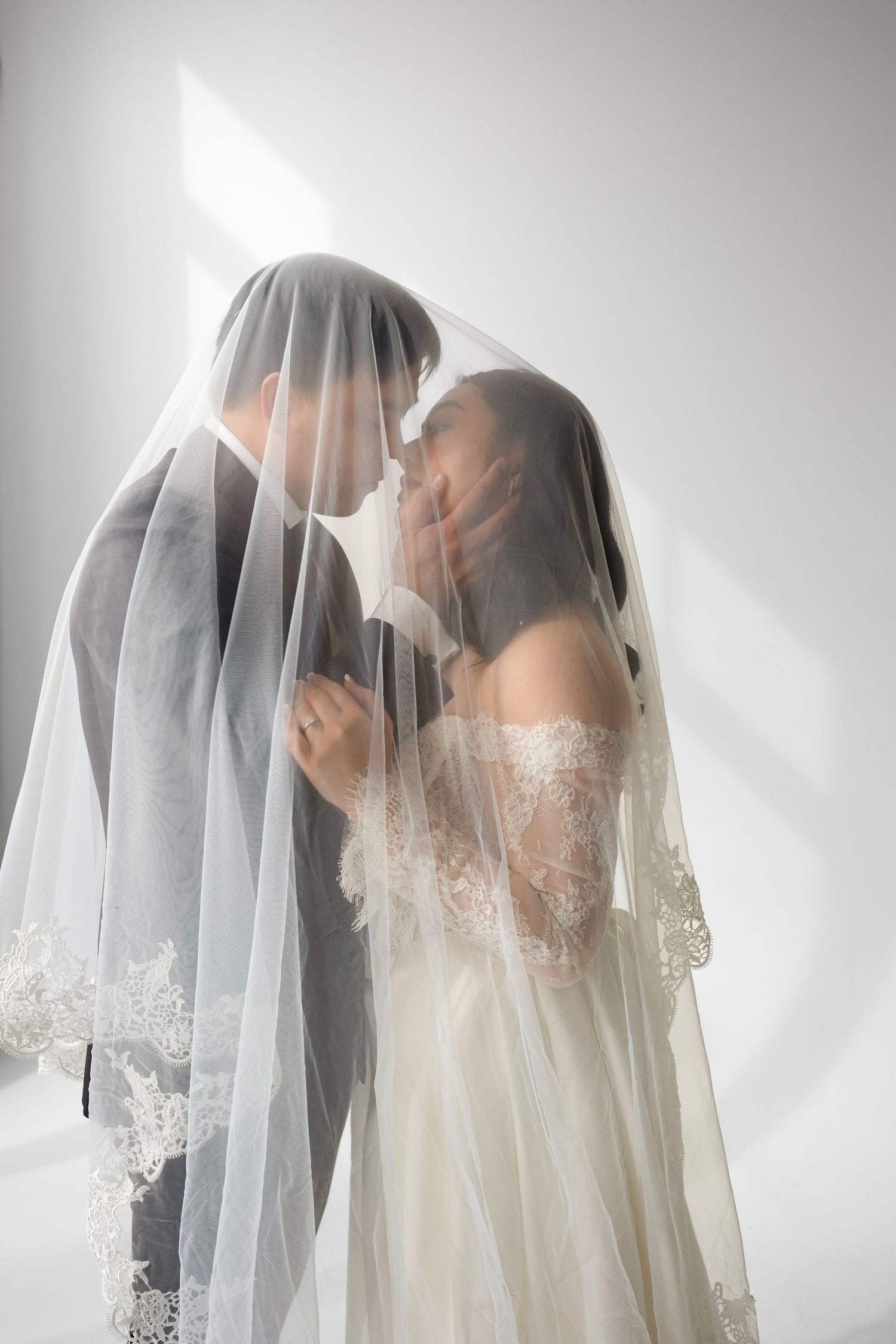 Bride and groom sharing a romantic kiss under a lace-trimmed wedding veil in a softly lit studio.